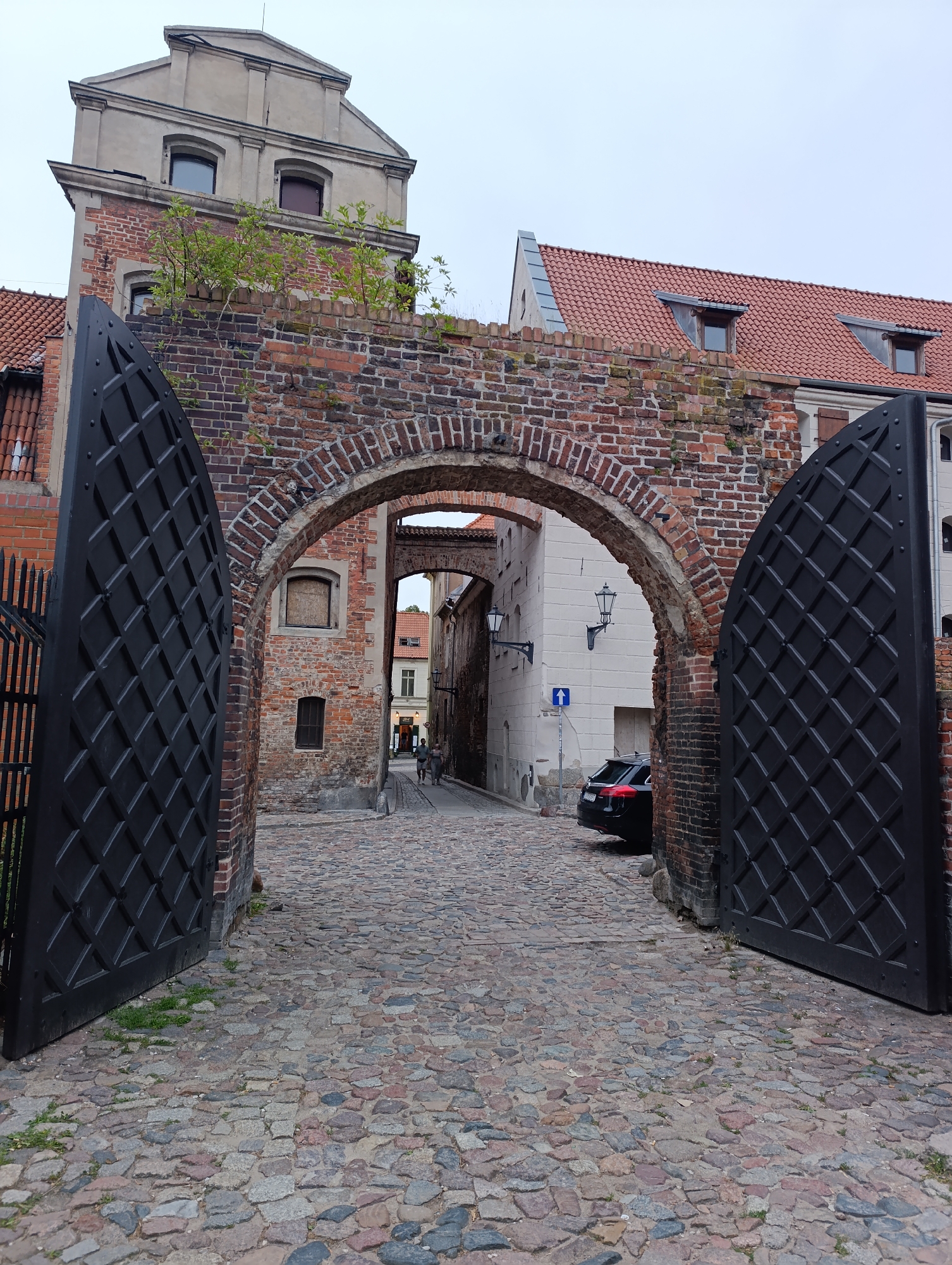 The image depicts a view through an open, ornate black gate made of metal. The gate is set within a brick archway, which appears to be part of an old city wall or building. Beyond the archway, a cobblestone street stretches into the distance, lined with historic brick and stucco buildings. The architecture suggests a European city with a medieval or old-town character.
On the left side of the image, a tall brick tower with a grey, gabled roof is visible. This structure has several windows and is adorned with greenery growing from its brickwork. To the right of the archway, red-tiled roofs of adjacent buildings are visible, with dormer windows.
Further down the street, two figures can be seen walking, and a black car is parked on the right side. A blue directional sign pointing upwards is also visible on the white wall of a building on the right. The sky is overcast and a pale grey.
This scene is located in Toruń, Poland, specifically at the Gate of the Holy Spirit (Brama Ducha Świętego).