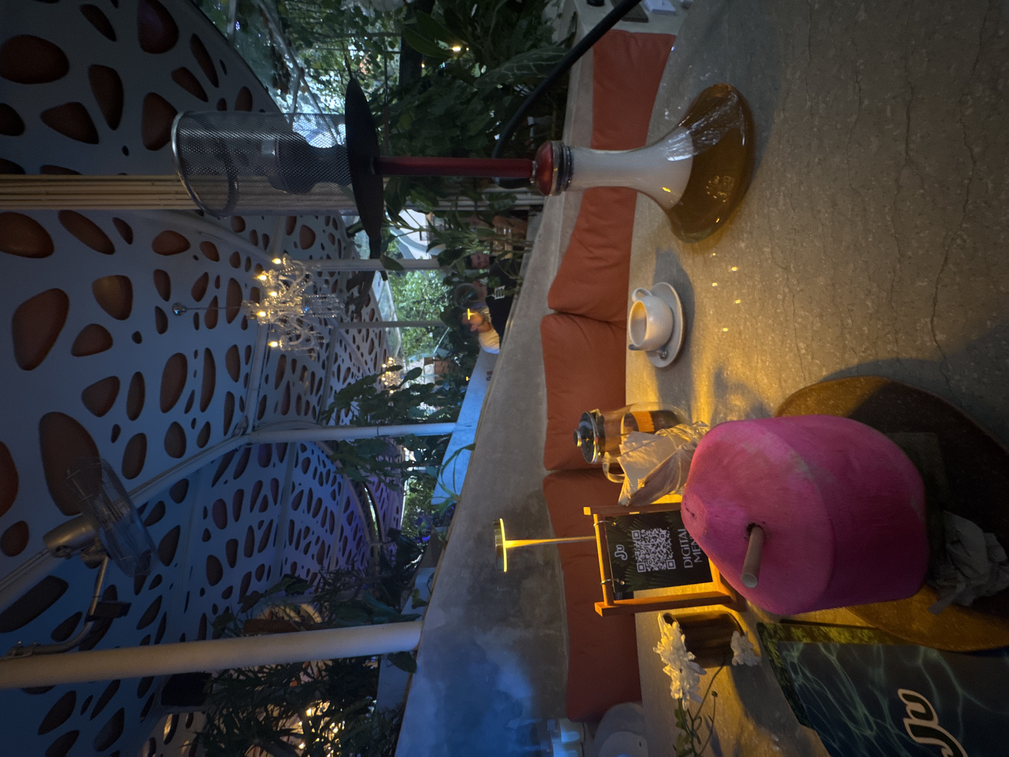 This image depicts an atmospheric outdoor or semi-outdoor lounge area at night, likely in North Kuta, Indonesia. The scene is oriented vertically in the provided image but can be interpreted as a horizontal view of a seating arrangement.
In the foreground and midground, a light-colored, textured table holds several items. A prominent hookah (shisha pipe) with a red stem and a white base, containing an amber liquid, is positioned on the table. Beside it, a small white teacup rests on a saucer. The seating consists of comfortable orange cushions along a built-in bench.
Other objects on the table include a glass teapot or infuser, a small table lamp with a golden stem, and a black-framed sign displaying a QR code. The sign clearly reads "DIGITAL MENU," indicating a modern ordering system. A notable pink, roughly cylindrical object with a stick or straw protruding from it is also present, possibly a beverage. Artificial white flowers and a glimpse of a dark blue surface with partial text "a la" are visible further down.
The background reveals a lush green environment with various plants and foliage. Overhead, the structure features distinctive white panels with organic, perforated patterns, through which a darker, possibly orange or brown, material is visible. Decorative light fixtures resembling small chandeliers with numerous tiny bulbs hang from the ceiling, casting a warm glow. A metallic mesh fan is attached to an overhead beam. A person can be vaguely seen seated in the background amidst the greenery. The time of day is clearly night, characterized by artificial lighting, which contributes to a relaxed and inviting ambiance.