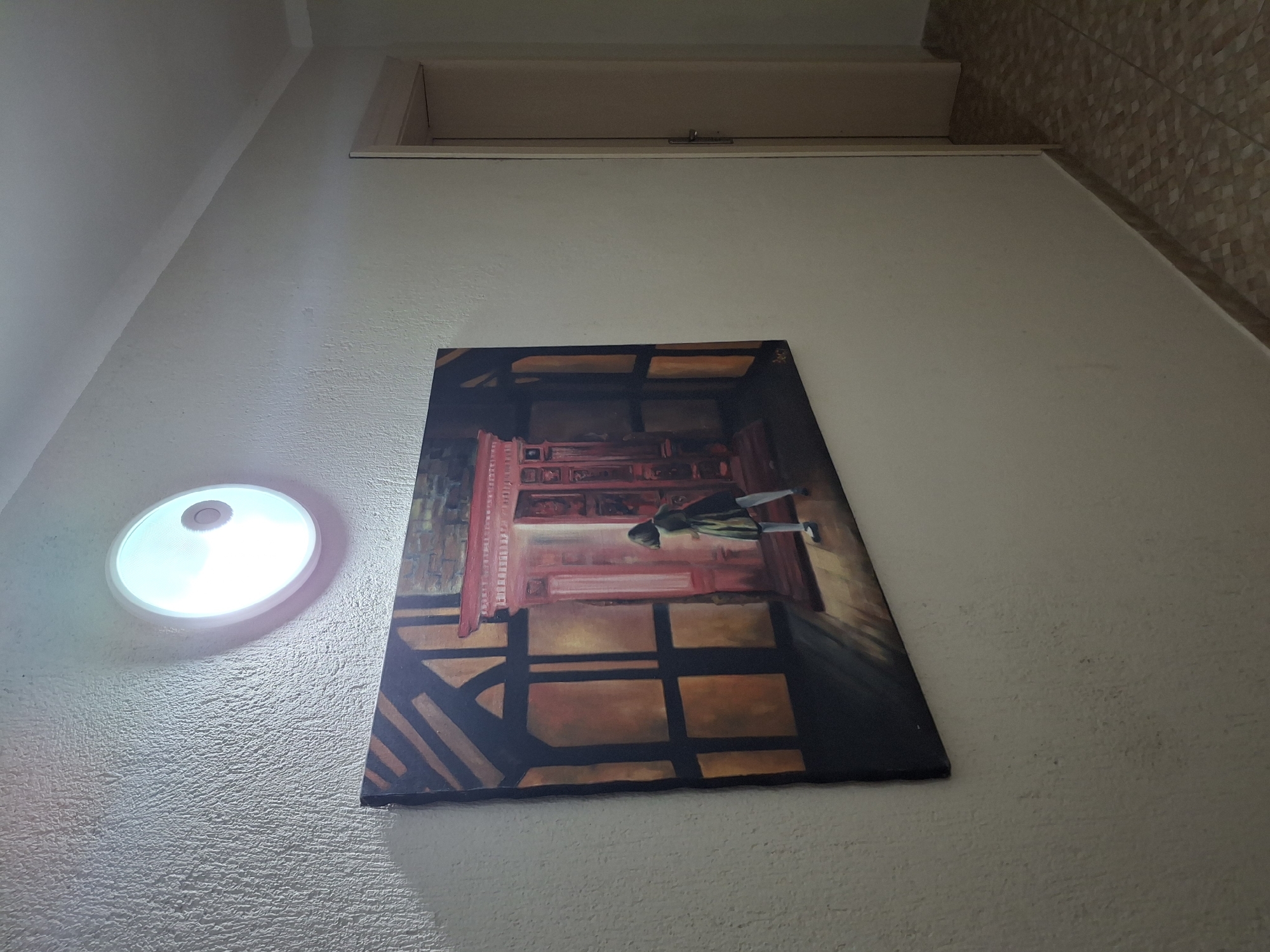 An eye-level, slightly low-angle, indoor shot shows a painting on a wall near a ceiling fixture, possibly in an apartment in Krumovgrad, Bulgaria. The wall is textured and painted light beige. The round ceiling fixture has a white plastic diffuser. Above the painting is a recessed closet or storage area, the bottom of which is flush with the wall. The painting is a rectangular canvas depicting an indoor scene, possibly an interior with a person. The painting's style is somewhat impressionistic, with warm colors dominating the composition. The perspective is slightly skewed, as if the photo was taken at an angle to the wall. The ceiling on the upper-right of the frame transitions into a wall with light beige tiles.