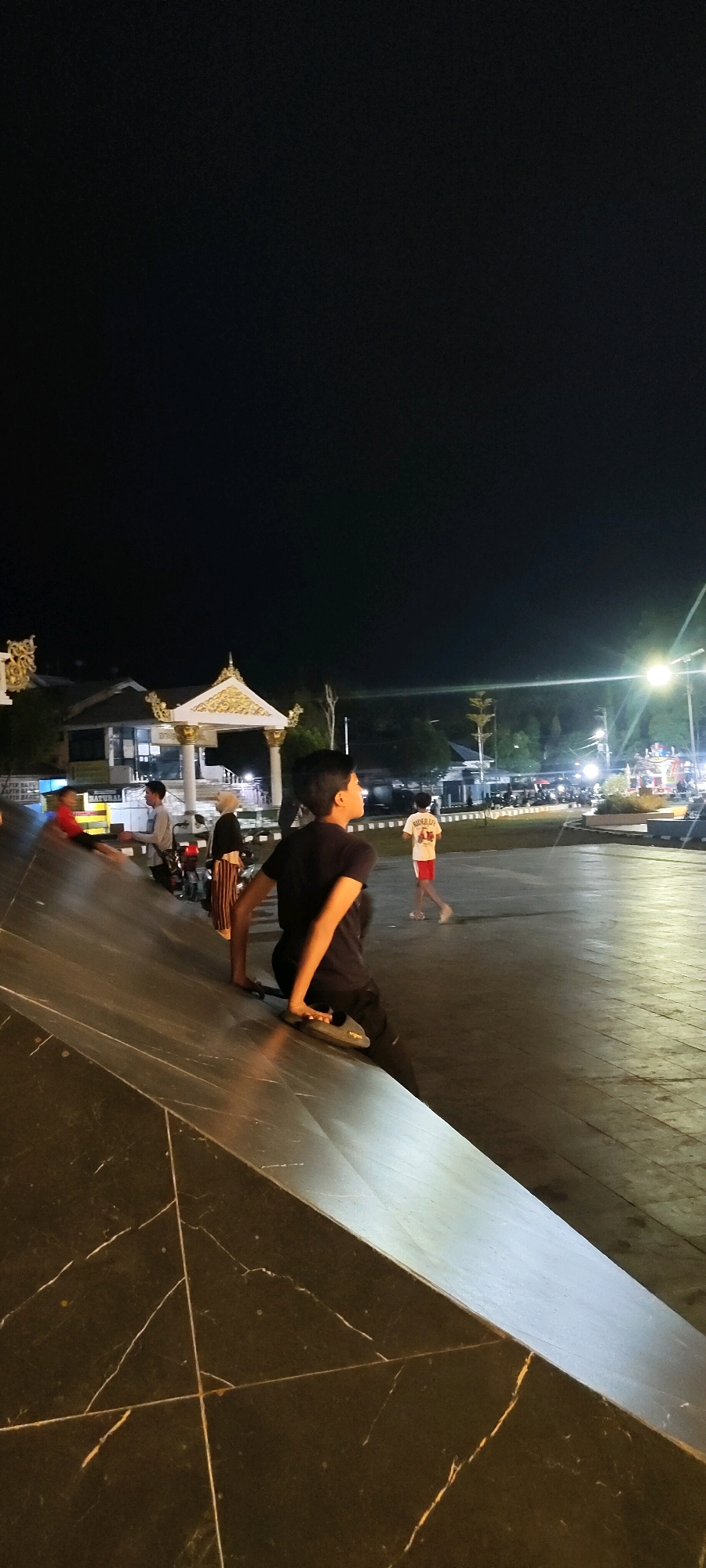 At night, in a public square in Martapura, Indonesia, a young man in a black t-shirt and black shorts sits on a slanted marble surface, facing away from the camera. His right leg is bent, and he appears to be resting his foot on a skateboard. His left hand rests on the skateboard. In the background, a group of people are visible. One person in a red shirt is on the slanted surface to the left. Further back, several individuals are standing near a covered structure with ornate golden trim. To the right, a person in a white t-shirt with red lettering and white shorts walks away from the camera, while another person in a white t-shirt and dark shorts walks towards the right. Streetlights illuminate the area, casting a glow on the paved ground and a few trees. The sky is dark and clear.