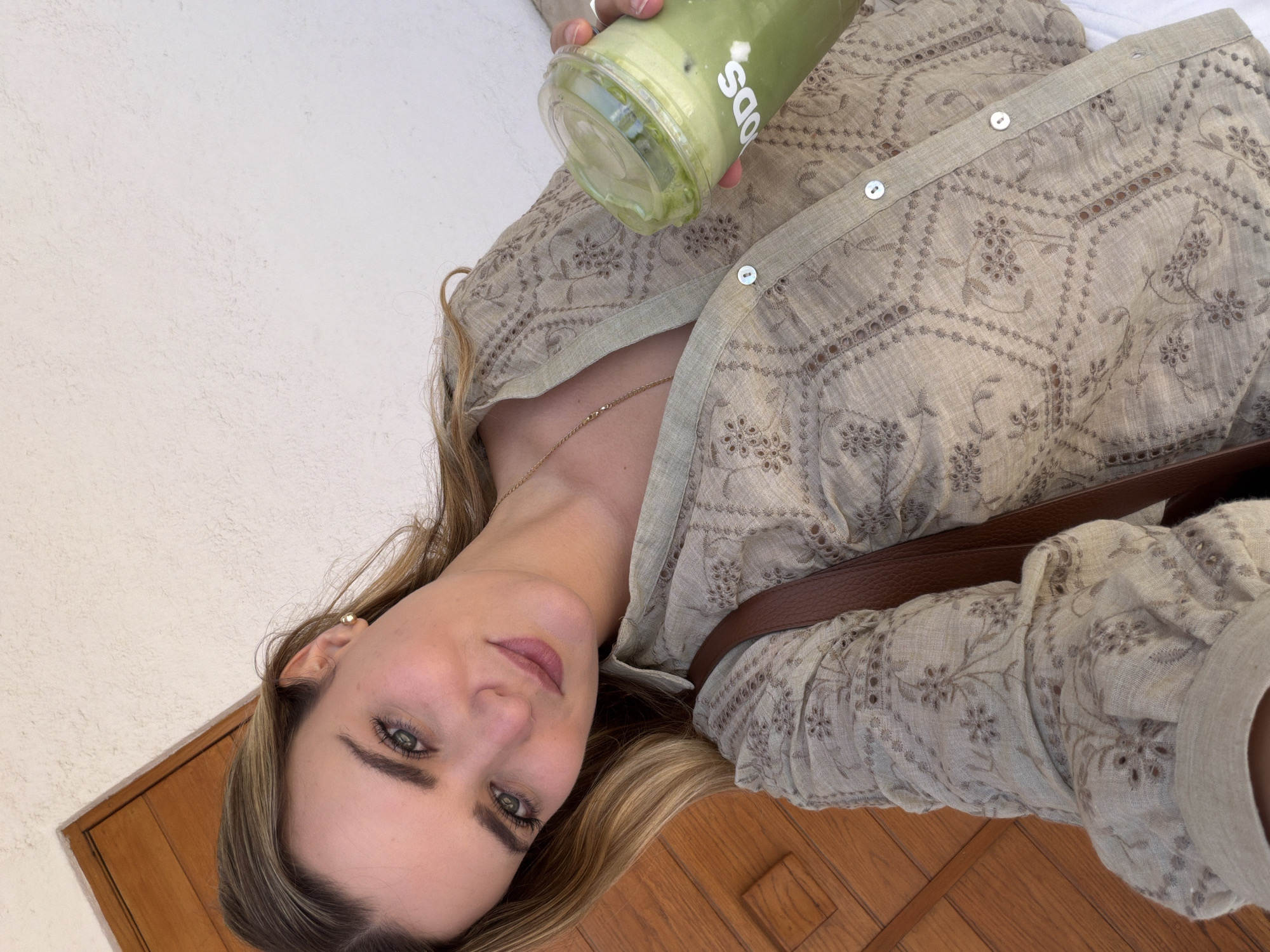 A woman with long blonde hair holds a green drink, possibly a smoothie, in a clear plastic cup with the label "ODS" visible. She is wearing a light beige, embroidered button-down shirt and a gold chain necklace. Part of a brown leather strap is visible across her body. She has light skin, green eyes, and is wearing makeup. The background consists of a light-colored, textured wall and a dark wooden surface. The location in the city of San Bernardino Tlaxcalancingo, Mexico is not discernible from the image.