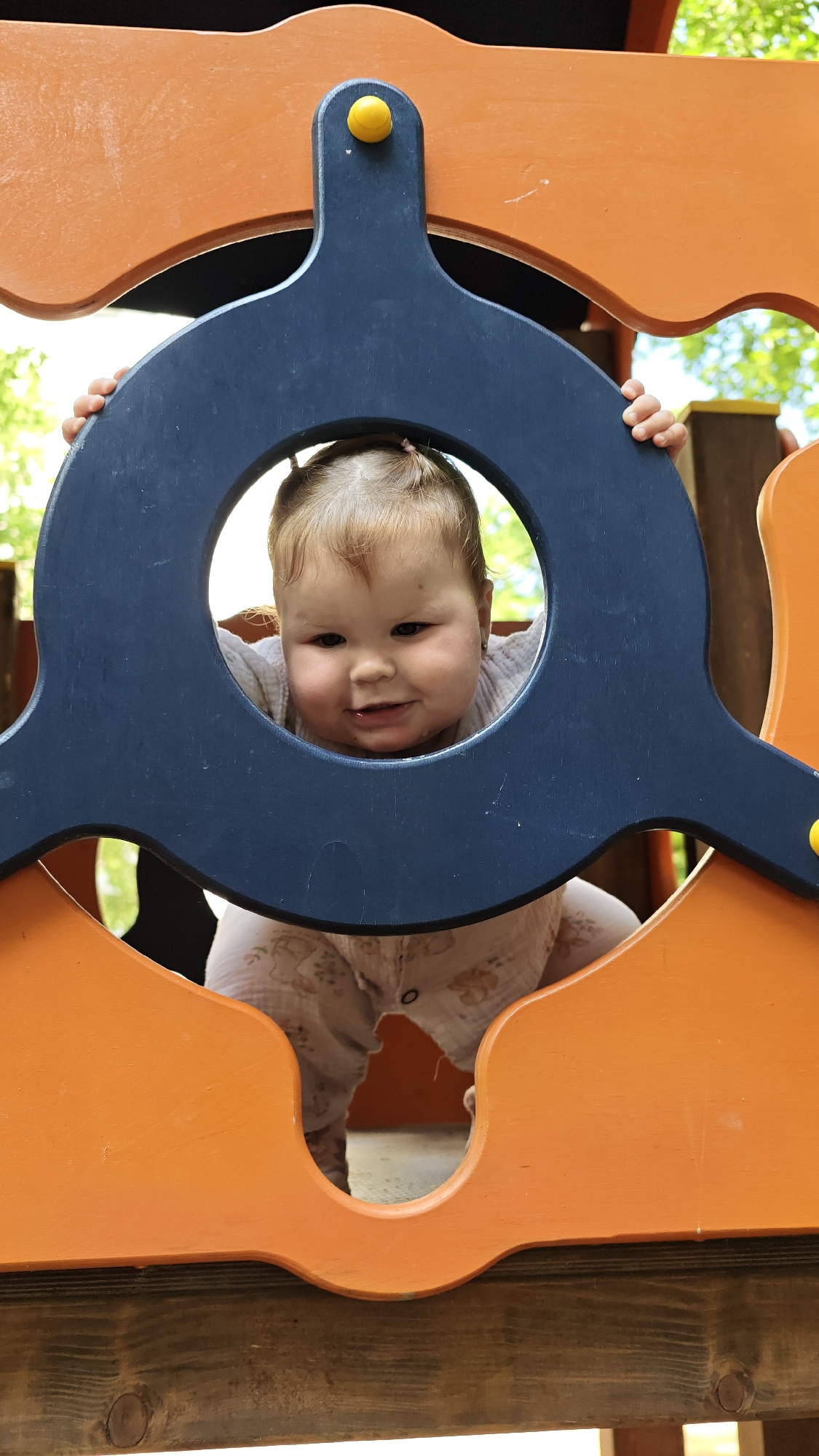 The image shows a baby peeking through a circular opening in a playground structure. The baby has fair skin, light brown hair pulled into two small pigtails, and is smiling. They are wearing a light-colored, patterned onesie. The playground structure appears to be made of orange and dark blue wooden elements, with a yellow knob visible at the top. The background shows blurred greenery, suggesting an outdoor setting. The image was taken at a playground in the city of Chaikovsky, Russia.