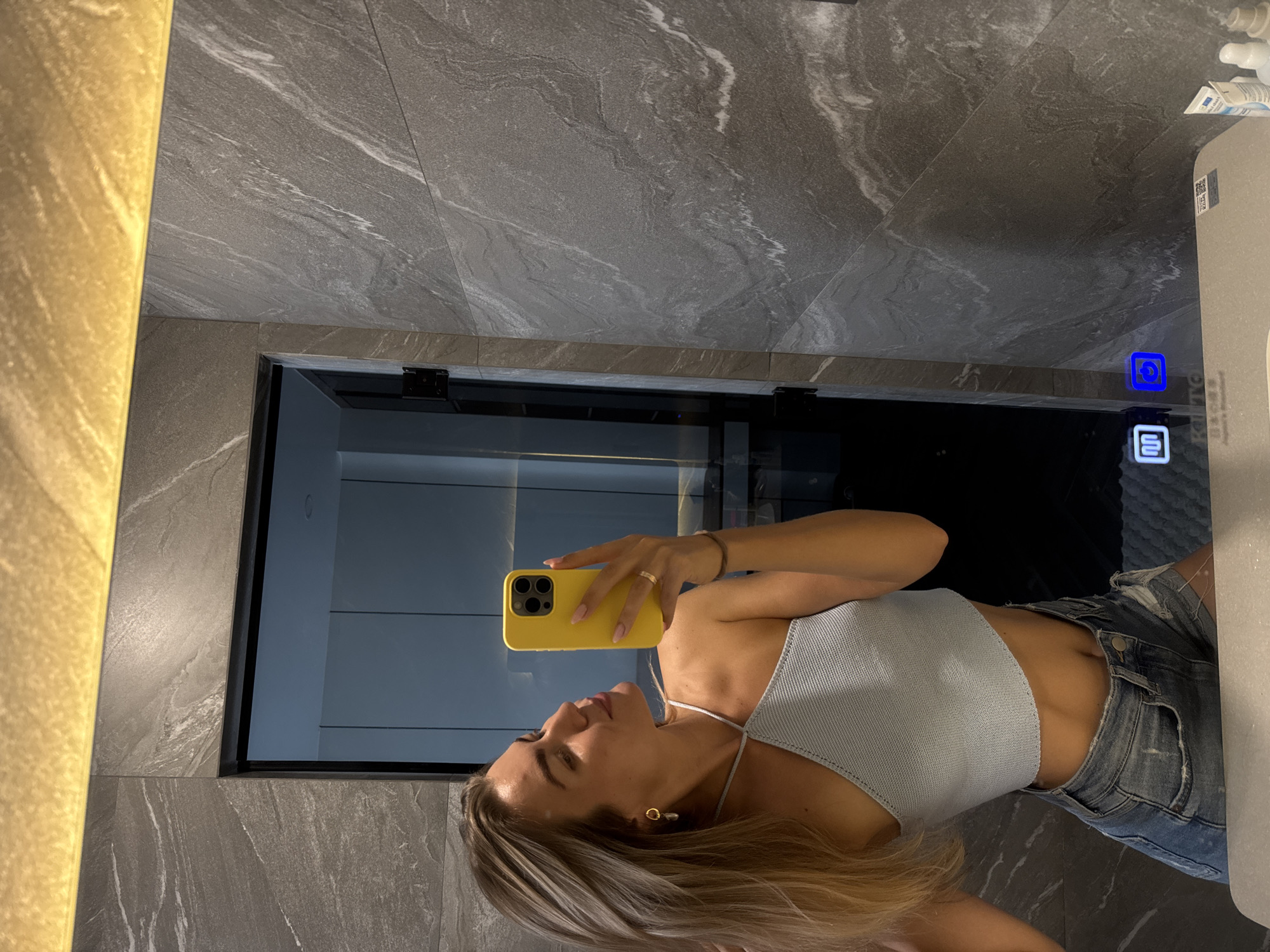 The image shows a person taking a selfie in a bathroom. The person is holding a yellow smartphone and is wearing a light blue top and denim shorts. The bathroom has gray marble walls and a mirror with an illuminated border. There are some toiletries visible on the right side of the mirror. The location is in the city of Unknown, Unknown.