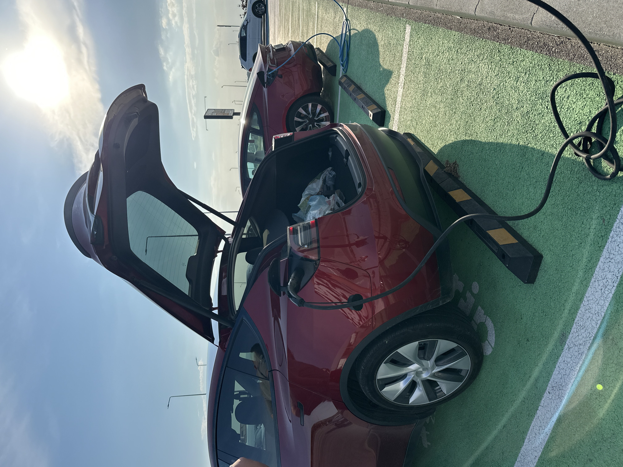 This forensic description details a daytime scene at a charging station in Kapitan Andreevo, Bulgaria. The image captures two red Tesla vehicles, both actively charging in designated green-painted parking spaces, under a bright sunlit sky with scattered clouds.
In the foreground, a dark red Tesla Model Y, recognizable by its open rear hatch and crossover body style, is parked with its rear facing the viewer. The trunk is open, revealing a plastic bag and a water bottle inside. A black charging cable is plugged into the vehicle's charging port, located on its rear left side, and extends down to the ground. Directly beneath the rear bumper, yellow and black striped wheel stops delineate the parking spot. The vehicle's silver multi-spoke wheel is visible on the left.
Behind the Model Y, a second red Tesla, a Model 3 sedan, is also parked and charging. A coiled blue charging cable is connected to its rear left charging port. Similar yellow and black striped wheel stops are present beneath its rear bumper.
The parking area features a distinctive light green, textured asphalt surface, marked with white lines defining the parking bays. Long shadows, indicative of a low sun angle (likely morning or late afternoon), are cast across the ground towards the viewer. In the mid-ground to the right, a white SUV is parked further away.
The background reveals a clear blue sky with some wispy white clouds. The sun, a bright luminous disc, is prominently visible in the upper right, contributing to the strong light and distinct shadows. Several dark, slender utility poles or streetlights are visible against the sky, along with a larger, dark rectangular sign structure in the distance on the right. No individuals are visible in the scene.