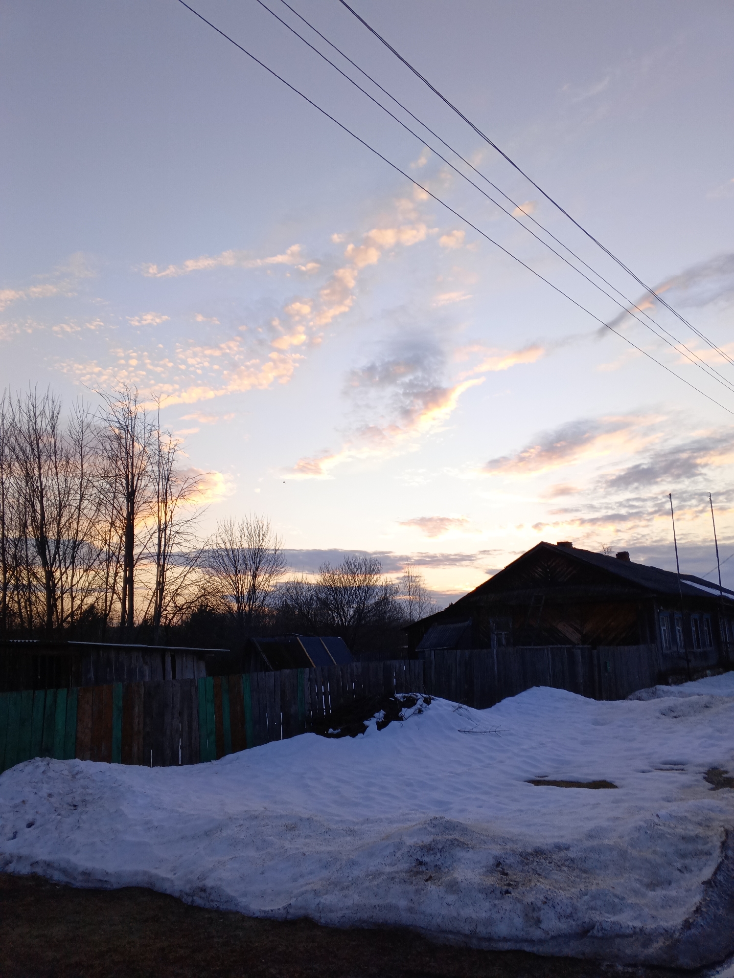 The image depicts a rural scene at dusk or dawn in Красный Маяк, Russia. The sky, occupying approximately two-thirds of the frame, transitions from pale blue-violet at the top to lighter hues near the horizon, featuring scattered cirrocumulus clouds illuminated by the setting or rising sun with faint orange, pink, and yellow tones. Four parallel power lines diagonally traverse the upper right quadrant of the sky.
In the midground, a substantial portion of the lower frame is covered by melting snowdrifts, exhibiting patches of white, blue-grey, and dirtier brown-grey where the snow is thin or mixed with earth. The immediate foreground appears as dark, wet ground. Behind the snow, a weathered wooden fence, composed of vertical planks in varying shades of green, brown, and grey, extends horizontally across the scene.
Behind the fence, on the left, a cluster of tall, bare deciduous trees with visible branches stands silhouetted against the lighter horizon. To the right, a dark, two-story wooden house with a steeply pitched roof is partially silhouetted. Multiple rectangular windows are discernible on its facade, and architectural details suggest traditional construction. Another smaller, dark wooden structure or shed is visible behind the fence to the left of the house. The overall environment suggests late winter or early spring conditions, devoid of visible human presence or activity.