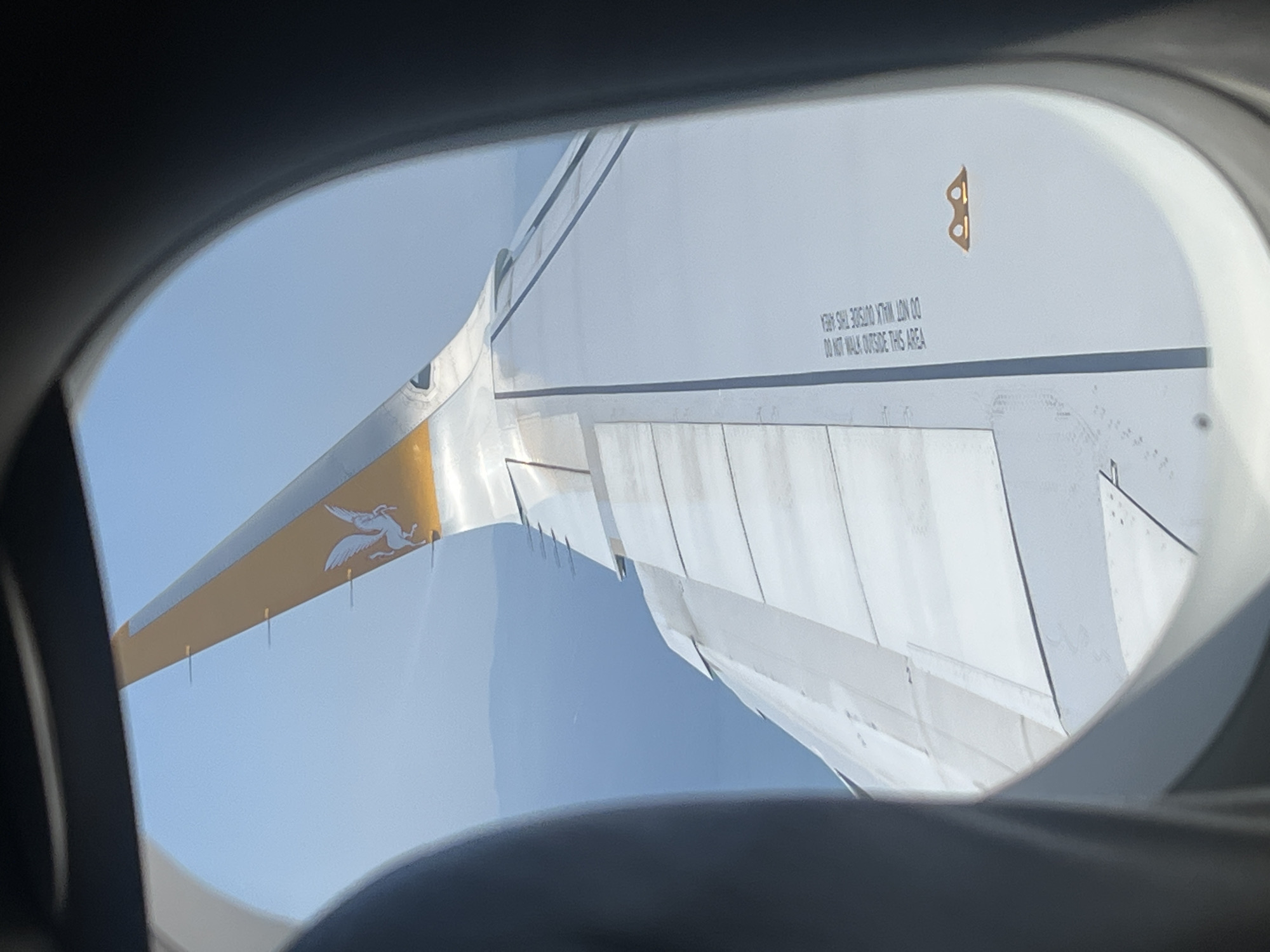 The image, taken from an airplane window, shows the wing of an aircraft against a clear blue sky. The wing is predominantly white with a golden-yellow tip. A white Pegasus logo is visible on the tail of the wing. The wing has several panel lines and some text that reads "DO NOT WALK OUTSIDE THIS AREA". A small symbol resembling a pair of goggles is also present on the wing. The photograph is taken in the city of Saint-Laurent-du-Var, France, as indicated by the metadata. The environment is that of an aerial view, with distant land masses visible beneath the wing. There are no people or interactions depicted in the image.