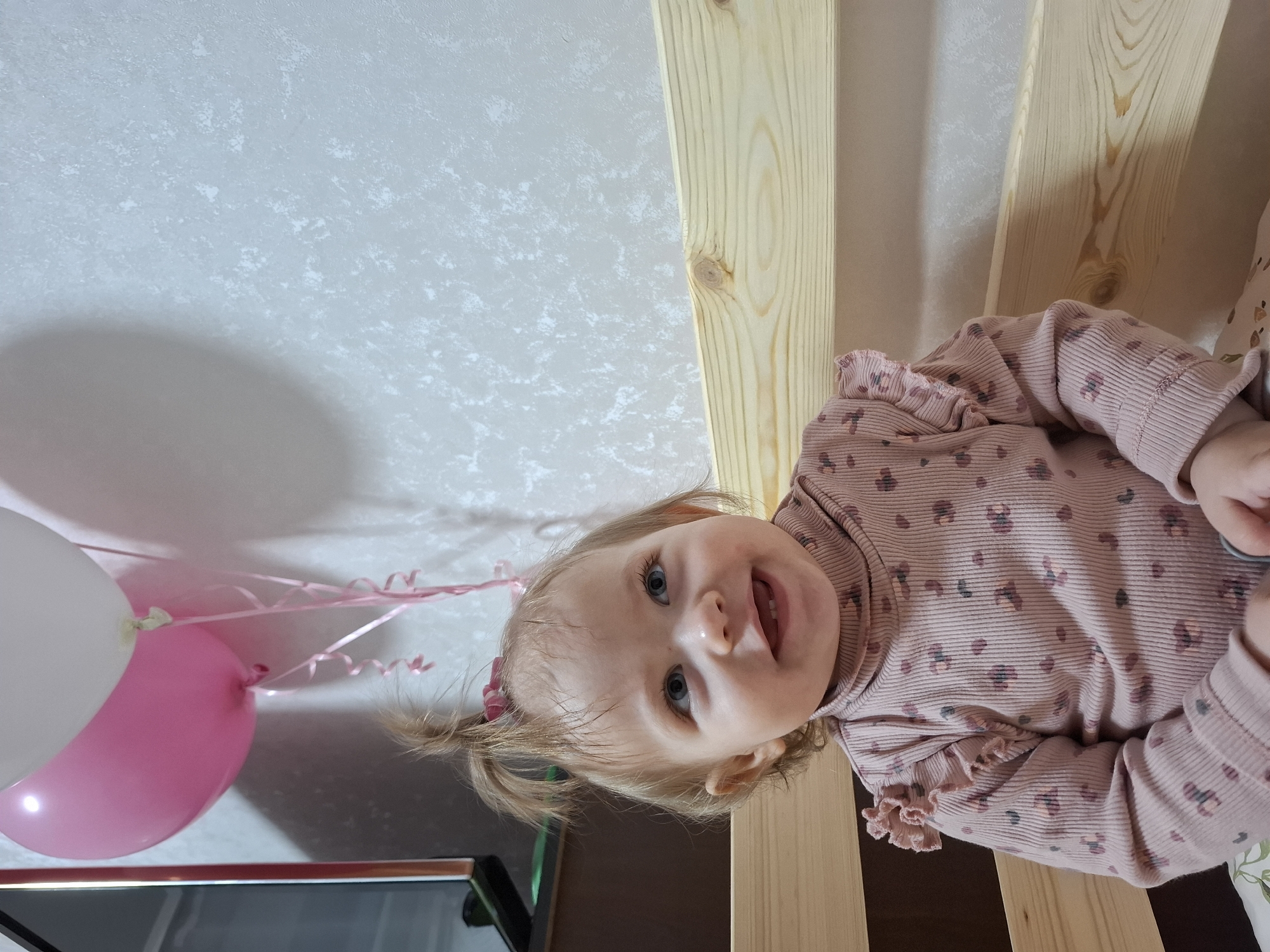 A young child with light blonde hair tied in small pigtails, wearing a pink floral-patterned long-sleeved shirt, is positioned in front of a light-colored wooden structure, possibly a crib or ladder. The child is looking directly at the camera with a slight smile. To the left, partially visible, are two balloons, one pink and one white, with curling pink ribbons attached. The background consists of a textured off-white wall. The setting is likely indoors. The city and location information is deduced from the context of the user's request history which could contain such information.