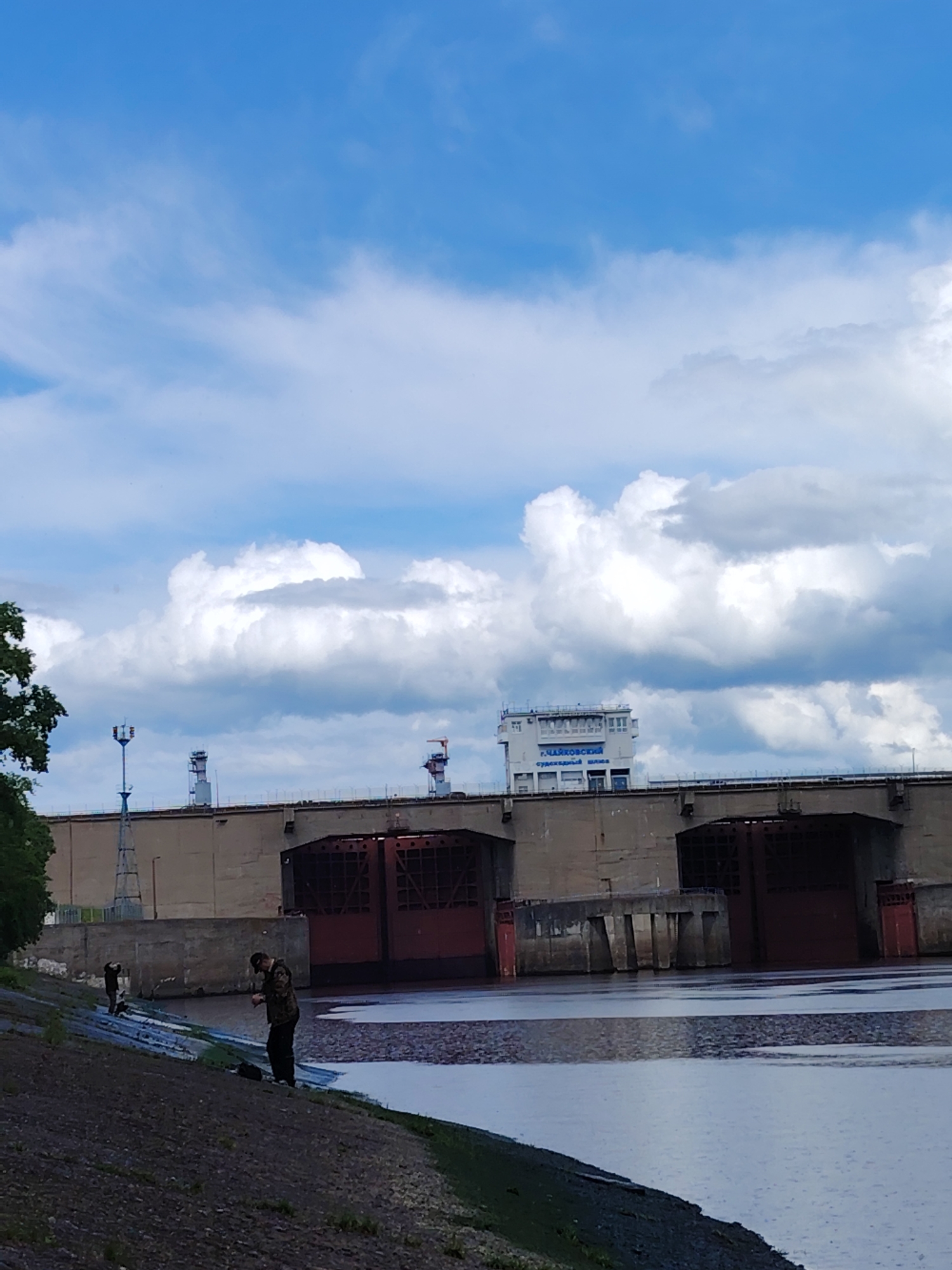 The image depicts a water dam in the city of Chaikovsky, Russia. Two people are standing on the grassy bank of a river, near the water's edge. The dam itself is a large concrete structure with red gates. Above the dam is a building with text in Russian, identifying it as part of the "Chaikovsky Ship Lock." The sky is partly cloudy, with a mix of blue and white clouds. Trees are visible on the left side of the image.
