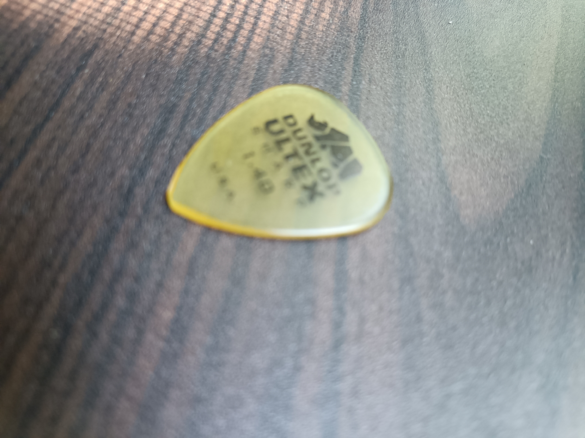 The image shows a guitar pick with the text "DUNLOP ULTE X 1.40 USA" on it. The pick is light yellow and has a triangular shape. It is lying on a dark, textured surface, possibly a wooden table. The background is blurred, with diagonal lines of light and shadow. There are no people or other objects visible in the image. The location in the city of Donetsk, Ukraine is not specified or deducible from the image.