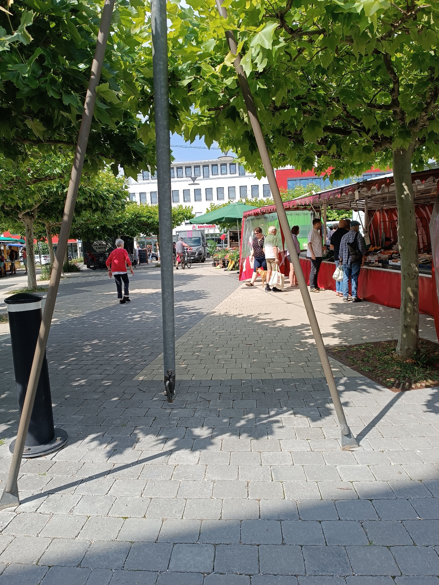 This image depicts an outdoor market scene in Germering, Germany, likely on a sunny day. The foreground is dominated by a paved walkway with a repeating brick pattern, dappled with the shadows of trees. Two large, metal poles, part of a temporary structure, extend vertically through the frame. To the left, a black trash receptacle with a white rim is visible.
In the middle ground, several people are browsing at market stalls. The stalls are covered with red and white striped awnings. One stall appears to be selling produce or food items, with visible fruits and vegetables. A woman in a red sweater and dark pants is walking away from the camera on the paved walkway, while another person on a bicycle is seen in the background. Behind the market stalls, modern buildings, including a white building with numerous windows and a red-accented building, are visible. The overall environment suggests a lively, public space with commercial activity.