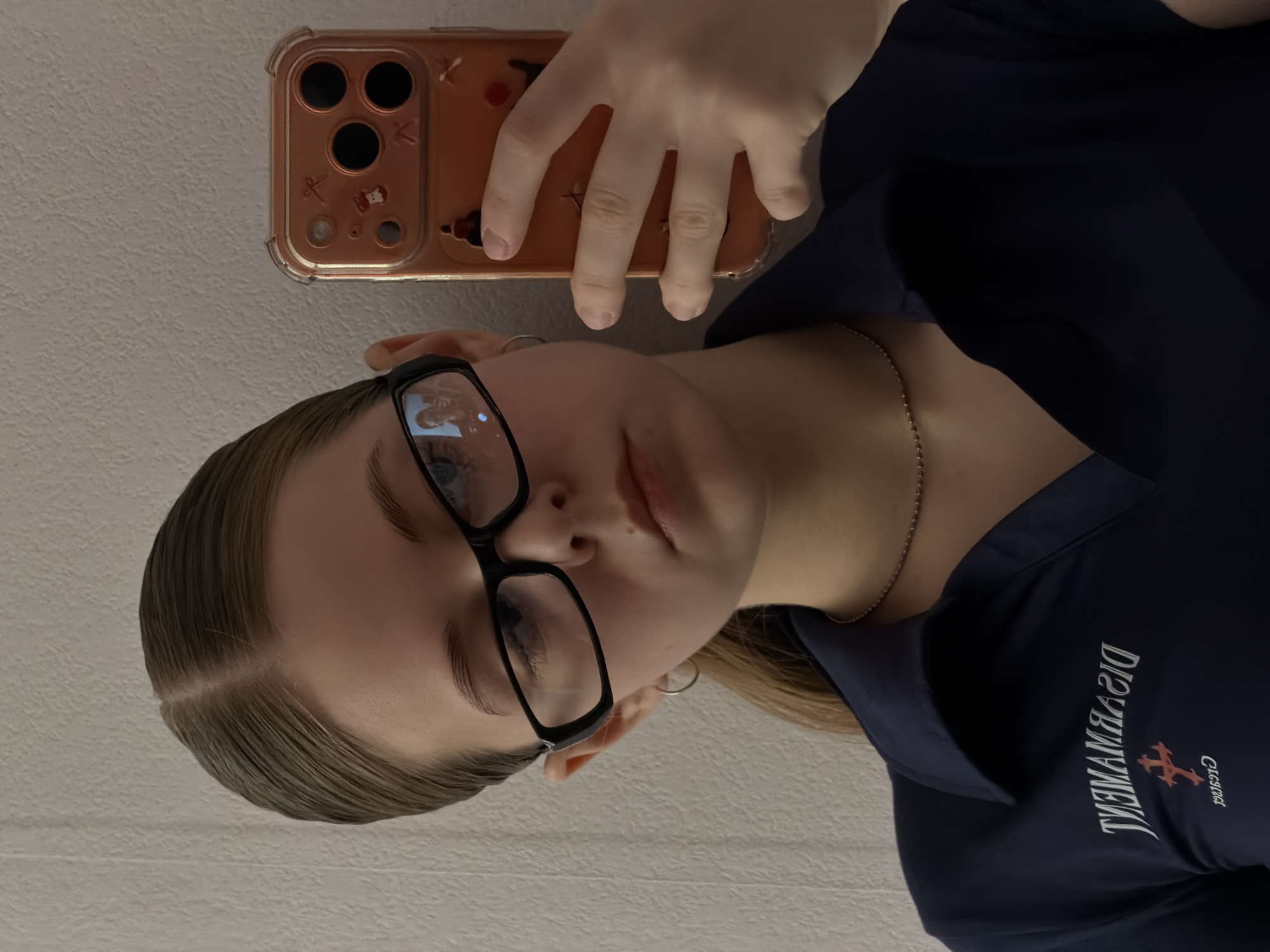 A selfie of a young Caucasian woman with light brown hair, wearing glasses and a dark blue polo shirt. She is holding a cell phone with an orange case in her right hand. The text "DISARMAMENT" is printed on her polo shirt, along with a logo. The background is a light-colored, textured wall. The city of Perm, Russia is not discernable from the image provided.