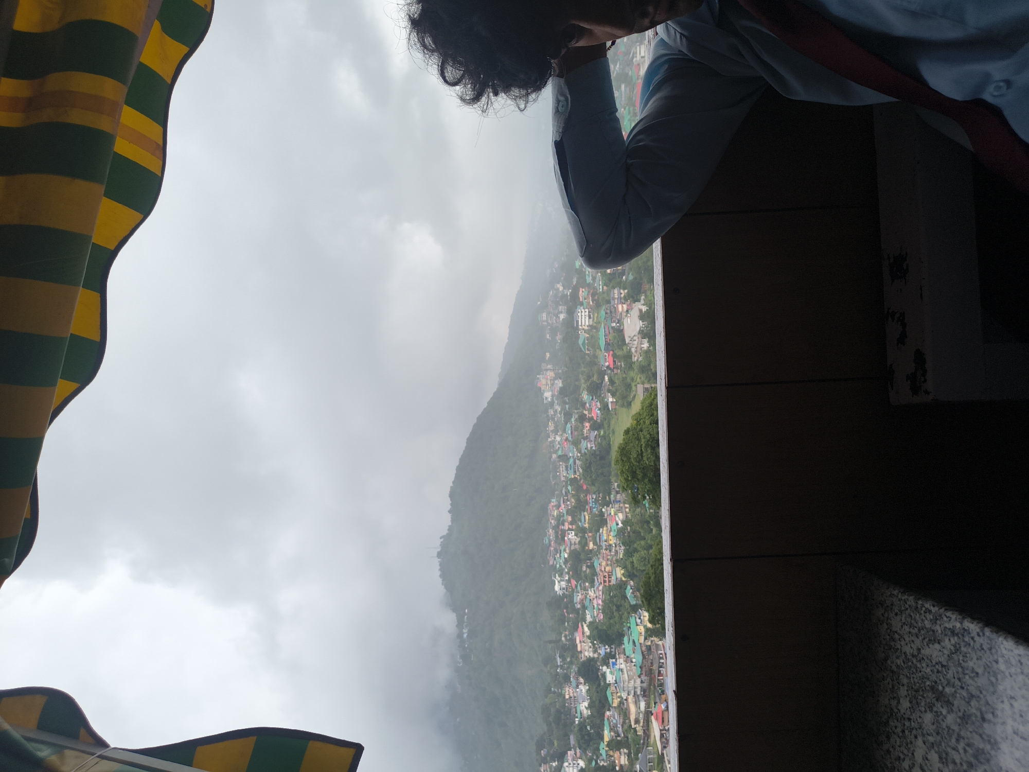 The image depicts a view of Dharamshala, India, on a cloudy day. In the foreground, a person with curly dark hair is leaning back against a railing, their right arm resting on their head. They are wearing a light blue collared shirt and a red tie. The background shows a densely populated hillside town with buildings of various colors, many with green roofs, nestled amidst lush green hills. A mountain range, partially obscured by clouds, rises behind the town. An awning with yellow and green stripes is visible at the top of the frame, suggesting the vantage point is from a covered area, possibly a cafe or hotel balcony. The overall atmosphere is overcast and slightly humid, typical of the monsoon season in the region.
