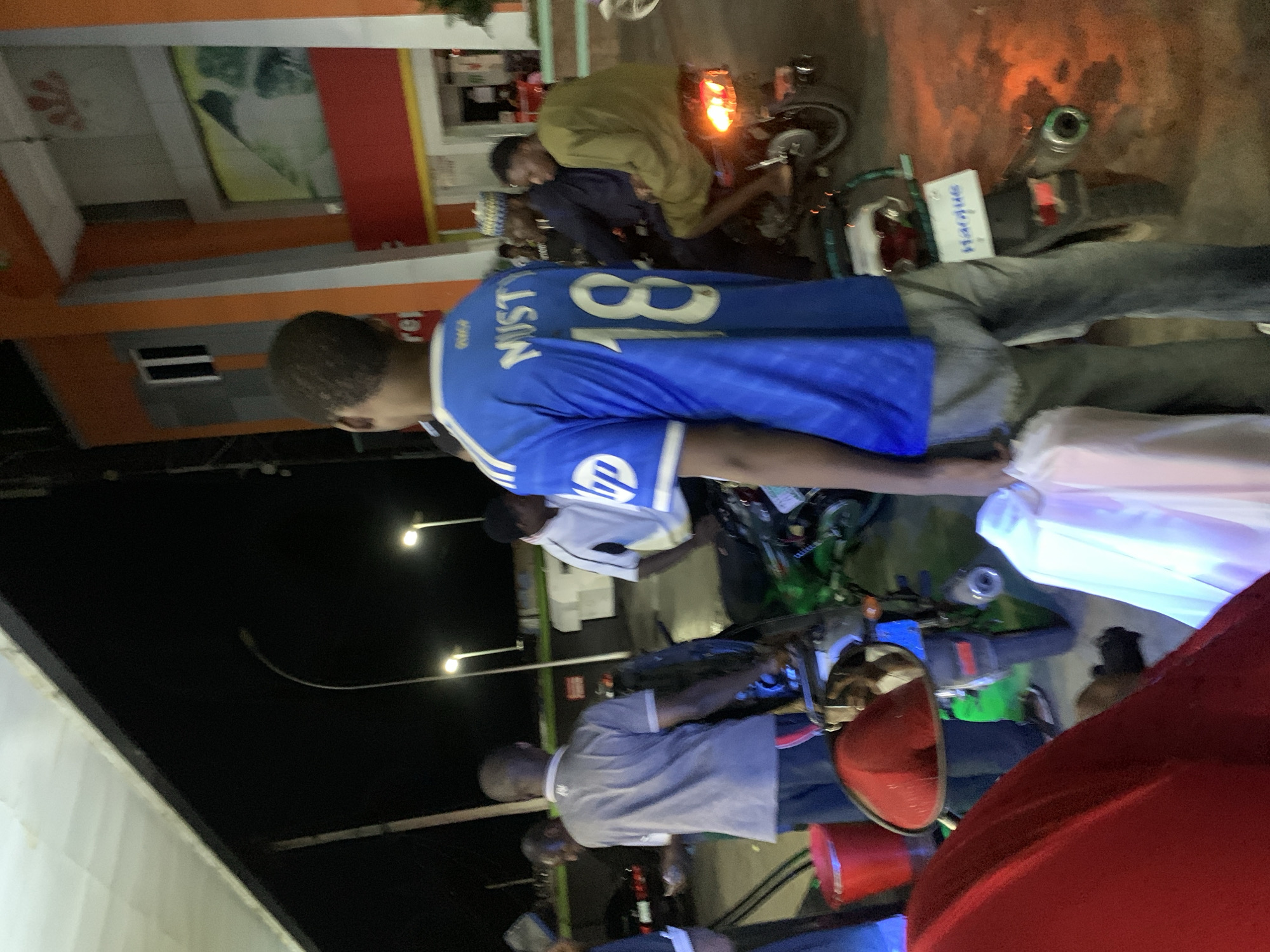 Here's a factual description of the scene:
The image shows a nighttime scene in Lokoja, Nigeria. The location is likely a roadside area with commercial buildings. There are several individuals present, engaged in various activities, many with motorcycles.
Specifically, one man wears a blue jersey with the number "18" and the name "Mustafa" on it. Two men are sitting on motorcycles; one is wearing an olive green top. There are other people standing or walking nearby. One man is wearing a grey shirt.
The background includes streetlights and buildings, some with visible signage. The overall impression is a busy street scene at night.