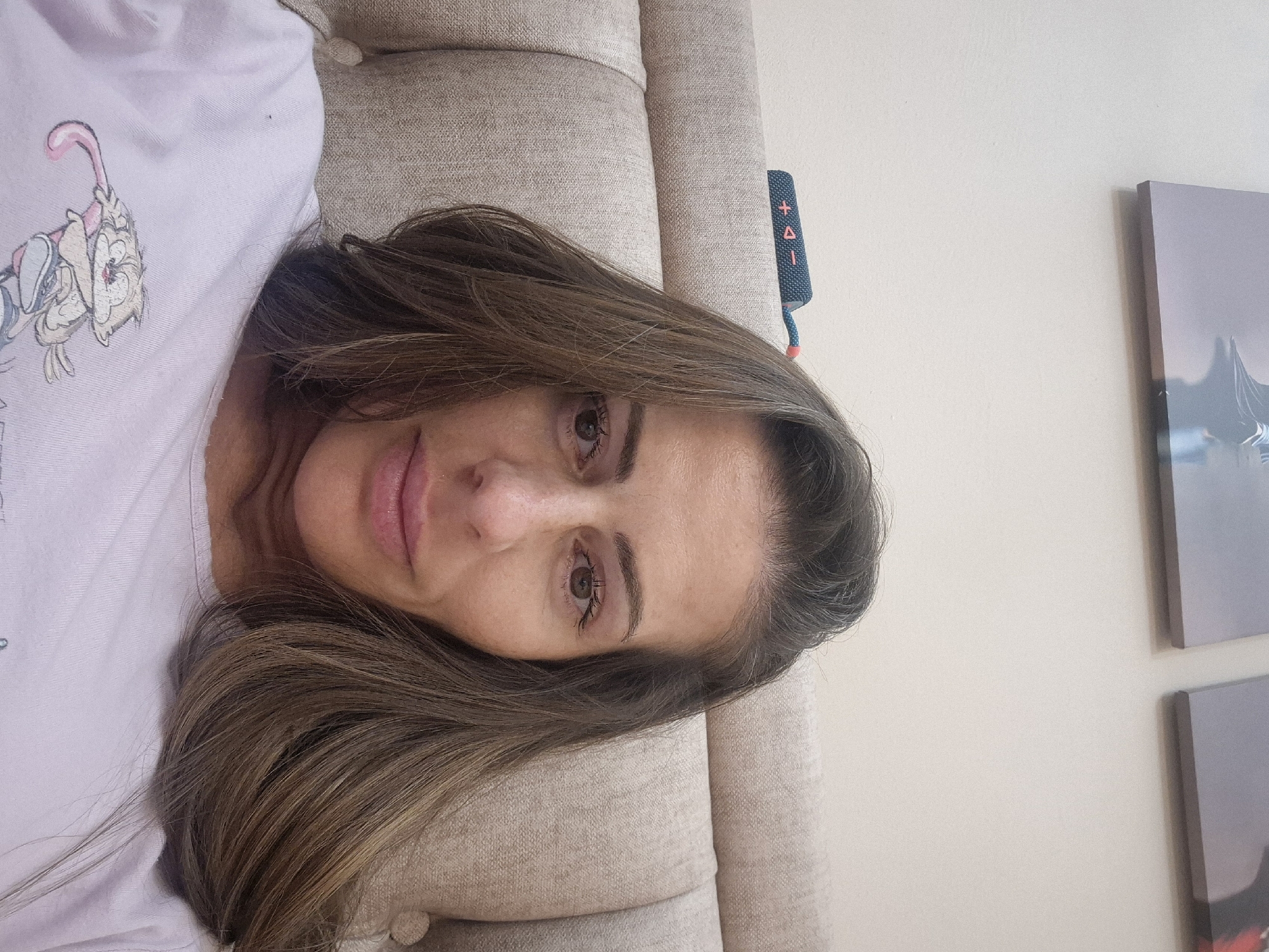 A woman with brown and graying hair is lying on a beige couch. She is wearing a light purple shirt with a cartoon graphic. A blue cylindrical object with orange buttons is on the couch near her head. Two framed pictures are hanging on the wall behind her. The location is an indoor setting in the city of Unknown, Unknown.