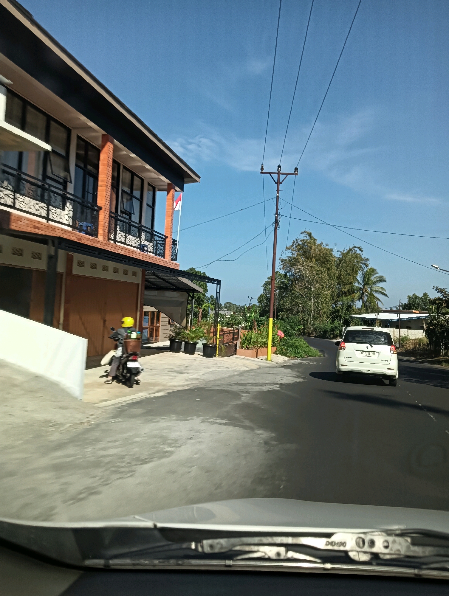 The image, captured from the perspective of a vehicle's interior, shows a street scene in Tomohon, Indonesia. On the left is a two-story building with red brick and dark trim, featuring balconies with black railings. A white wall curves away from the building in the foreground. A person wearing a yellow helmet rides a motorcycle in front of the building. To the right, a white car is driving away on a paved road. Tall trees and power lines are visible in the background under a clear blue sky. The foreground shows the vehicle's dashboard and windshield wipers.
