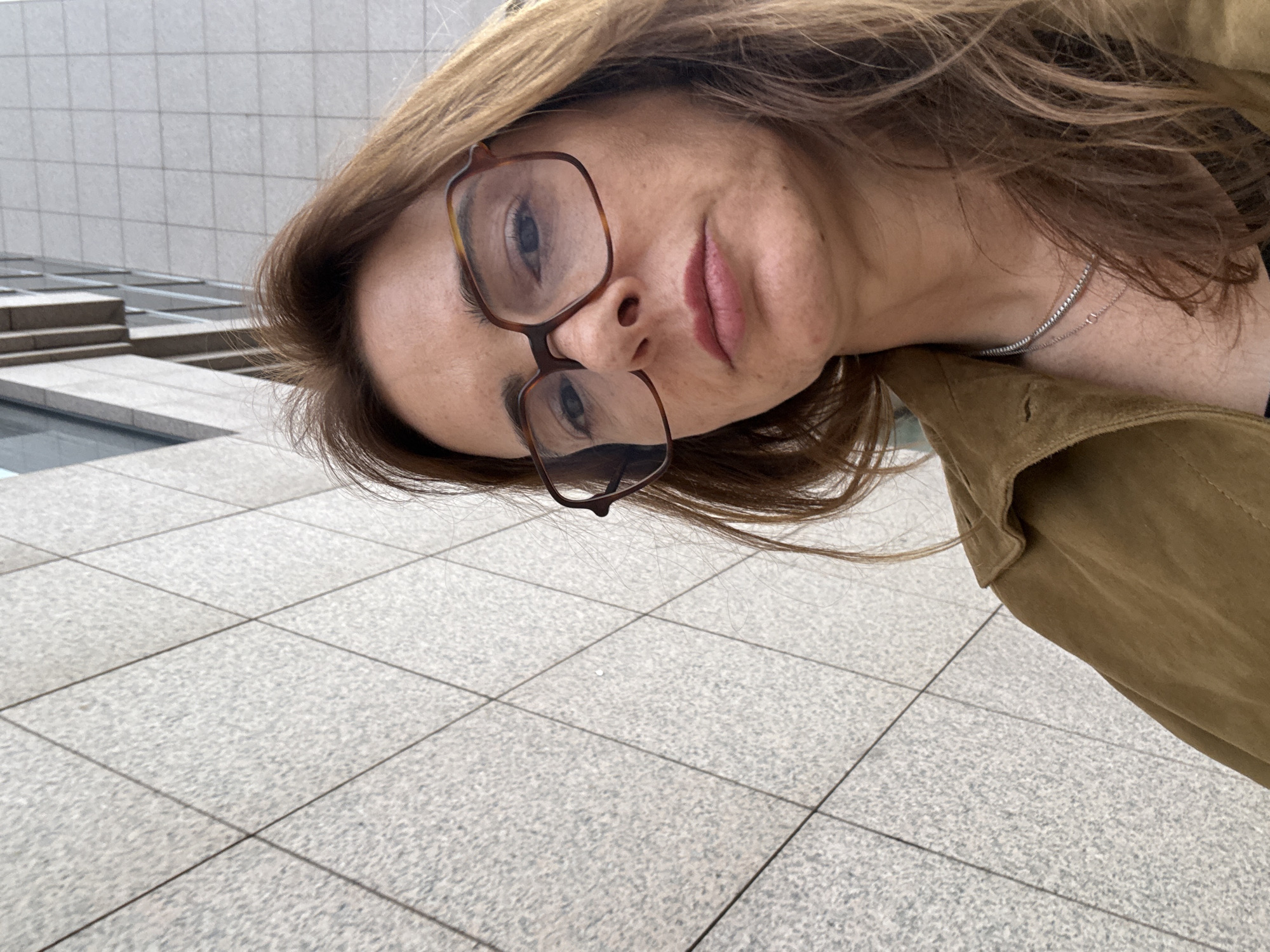 A tilted selfie depicts a fair-skinned woman with light brown hair wearing glasses and a tan-colored jacket. She is also wearing a delicate necklace. The background features a tiled surface and a building with a grid-like facade, suggesting an urban setting. The photograph appears to be taken in Lisbon, Portugal, as evidenced by the architectural style and the type of tiling commonly found in the city.