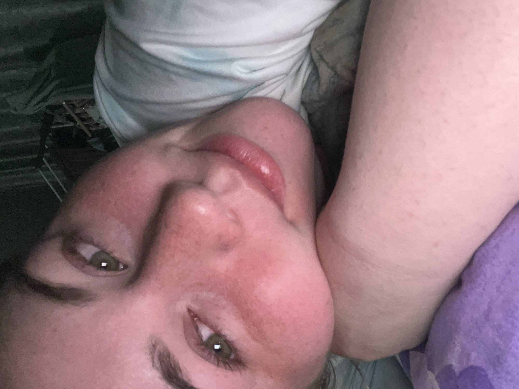A close-up, angled self-portrait of a person with fair skin, freckles, and light brown hair. They have hazel eyes and glossy lips. They appear to be lying down on a surface, possibly a bed. To the left of the person, there are dark objects. It is impossible to identify if this photo was taken in the city of Chaikovsky, Russia without further information.