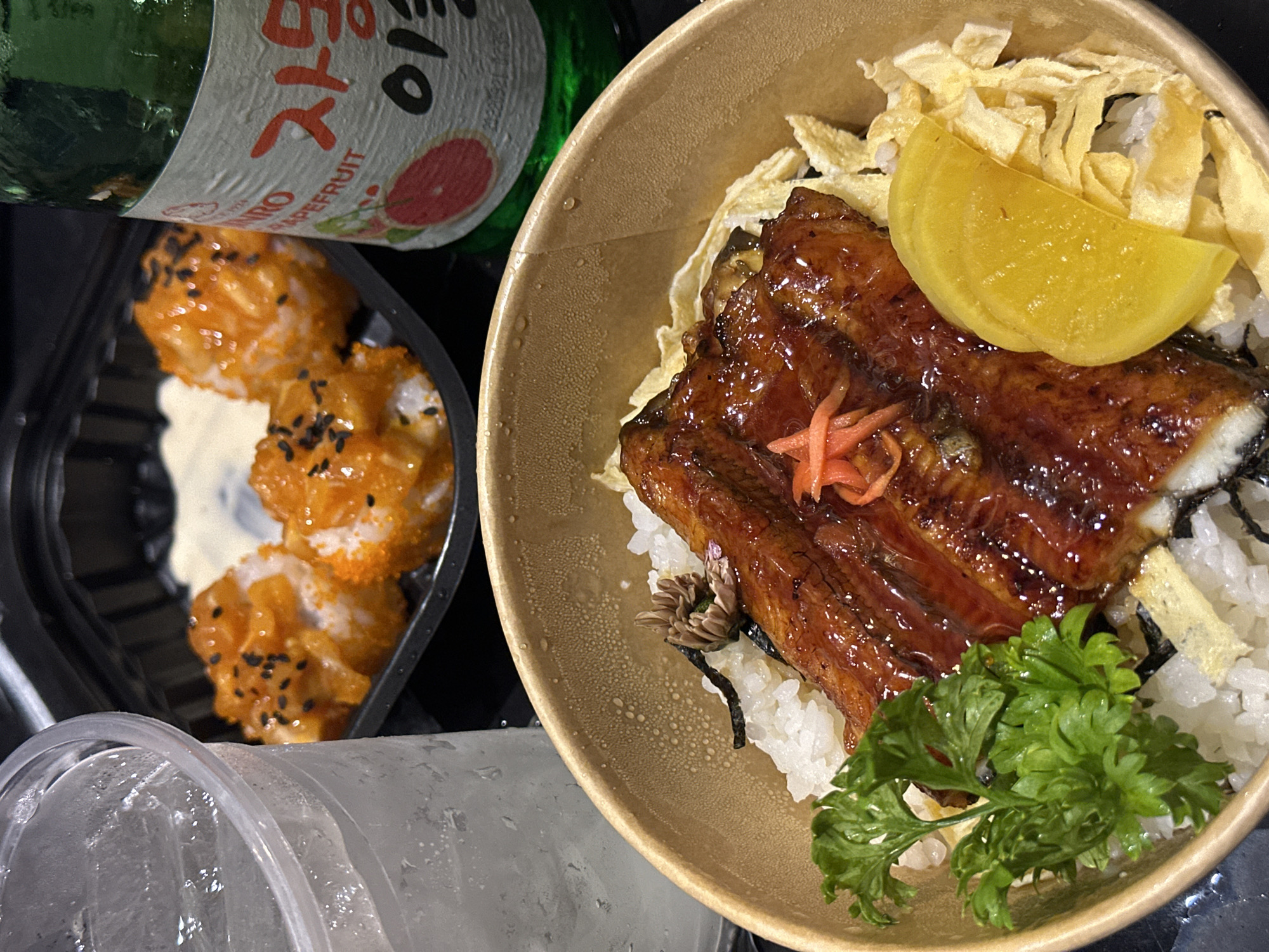 The image depicts a meal consisting of eel over rice, tamago, pickled radish, and a side of spicy tuna rolls. A partially visible bottle of Soju is also present. The setting appears to be indoors, likely a restaurant or private residence. The overall presentation and type of food suggest the location may be in Đồng Nai, Vietnam, given the popularity of Soju and Japanese-inspired cuisine in the region. The image lacks identifiable individuals or interactions. The focus remains on the food items.