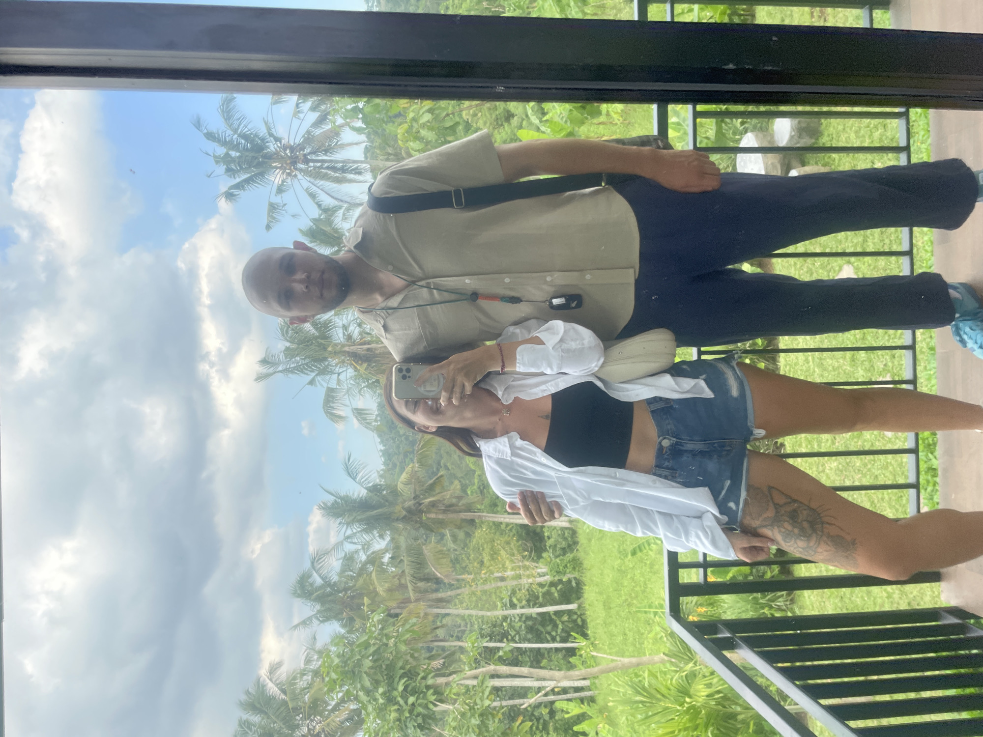A mirror selfie shows a couple standing on a balcony with a lush green jungle and palm trees in the background. The woman on the left wears a black crop top, a white open button-up shirt, and denim shorts. She has a cat tattoo on her left thigh. The man on the right is bald and wears a beige short-sleeved button-up shirt and dark pants. He has a black messenger bag slung across his shoulder. The woman holds a phone up to take the photo, and her arm is around the man's shoulder. The balcony has black metal railings. The sky is partly cloudy with blue patches. The location is in Penebel, Indonesia.