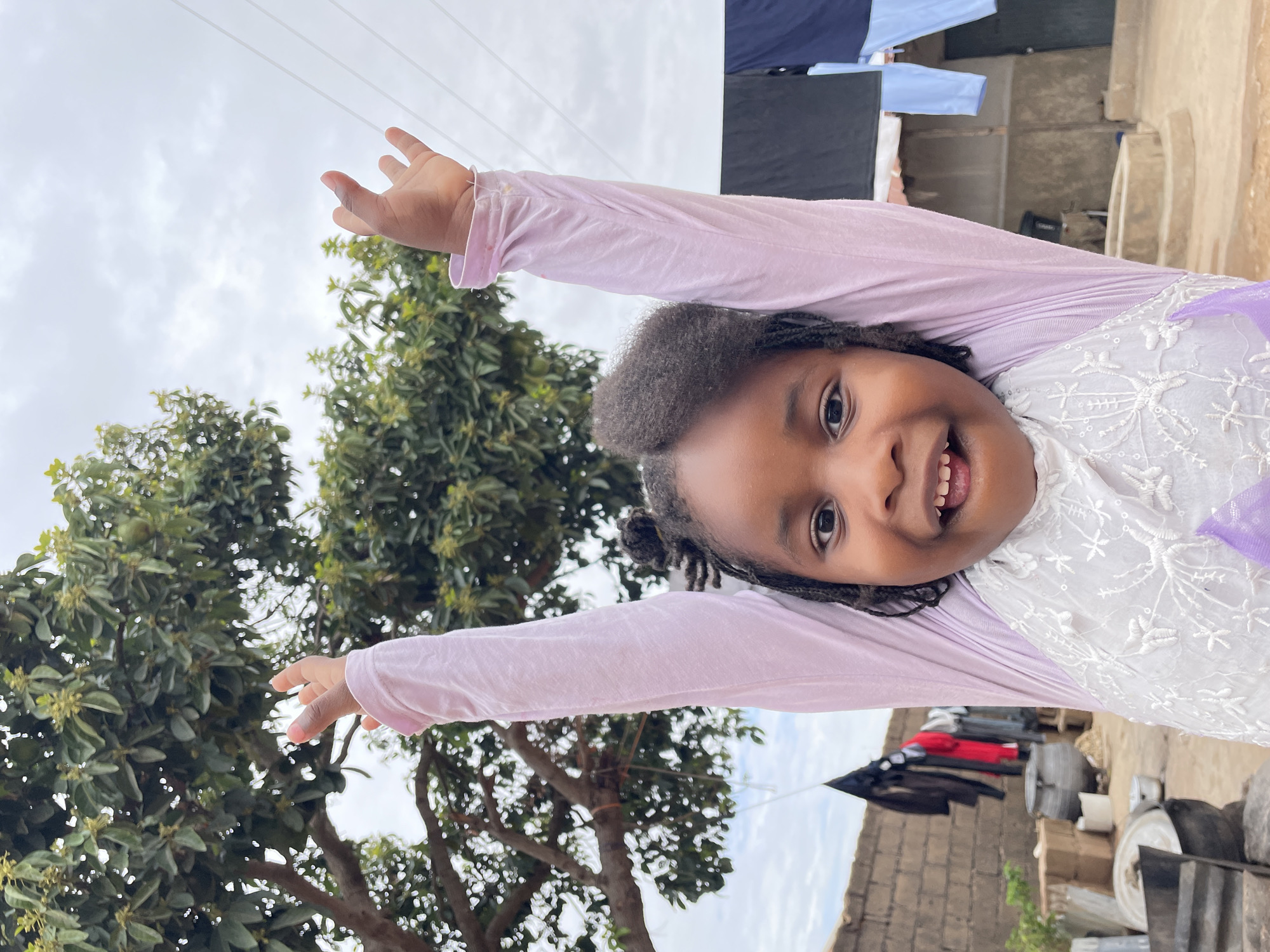 A young, dark-skinned girl with braided hair is smiling widely at the camera with her arms raised. She wears a light purple, long-sleeved shirt over a white dress with floral embroidery. Behind her is a tree with green foliage. In the background, there is laundry hanging on a line. A brick wall and various objects are also visible. The photograph appears to be taken outdoors in Jos, Nigeria.