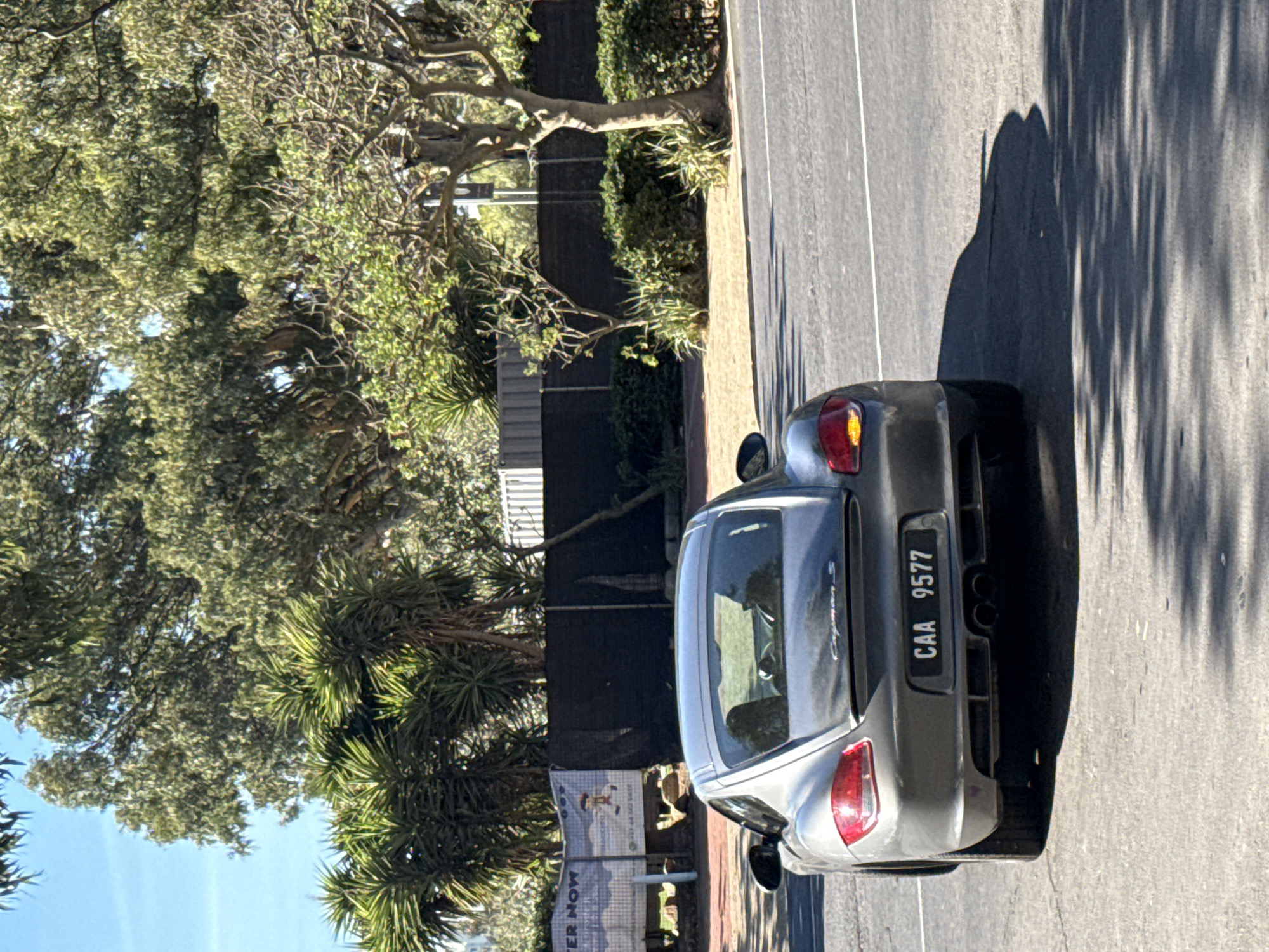 A gray Porsche Cayman S with license plate "CAA 9577" is parked on the side of a road in Cape Town, South Africa. The location appears to be a residential area with trees and a fence lining the roadside. A sign that reads "WATER NOW" is visible. The car is the primary subject. There are no people visible. The road surface is asphalt. Tree shadows are cast on the road surface.