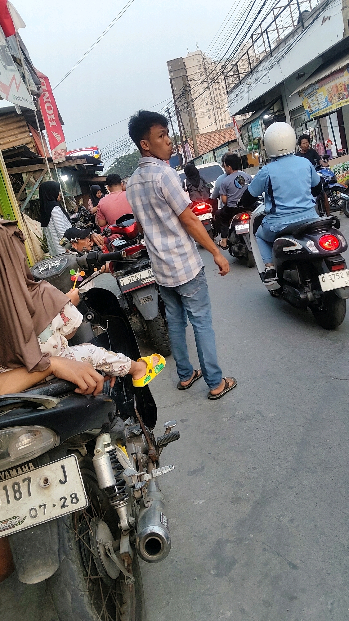 The image depicts a street scene in Mekarmukti, Indonesia, likely during daylight given the ambient light. In the foreground, a young man wearing a checkered shirt and blue jeans stands looking to his right, with a motorcycle partially visible in the bottom left corner. A child is seated on a motorcycle in the center-left, wearing a brown headscarf and light-colored overalls with cartoon characters, and bright yellow sandals. Other individuals are visible on motorcycles in the background, suggesting traffic or a gathering. The environment includes buildings with signage, power lines overhead, and a paved street. The overall atmosphere appears to be casual and possibly crowded, with multiple motorbikes present, common in urban Indonesian settings.