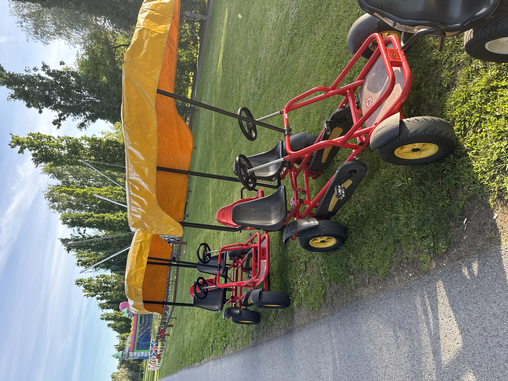 In Tallinn, Estonia, two red four-wheeled pedal go-karts, each equipped with a yellow canopy, are parked on a grassy area next to a paved pathway. The go-karts have black seats and steering wheels. In the background, several tall trees are visible, and further back, a large inflatable castle structure and trampoline equipment are present. The sky is blue with scattered clouds. The scene appears to be a park or recreational area.