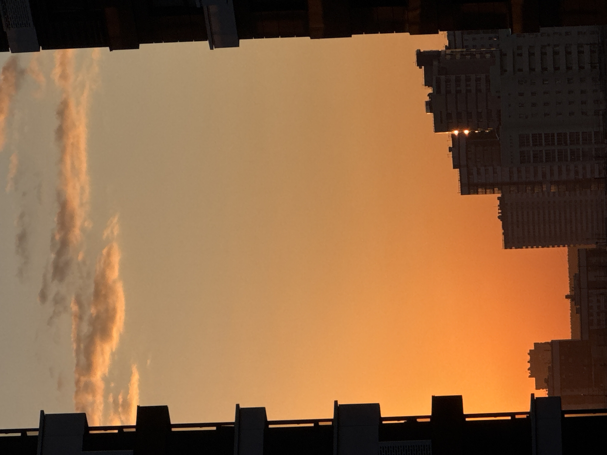 This image captures a vibrant sunset over the cityscape of Novosibirsk, Russia. The sky is dominated by warm hues of orange and yellow, with scattered clouds tinged in pink. Silhouetted against this colorful backdrop are several multi-story buildings, appearing dark and stark. On the left side of the frame, a portion of a dark building is prominently visible, with its balconies and structural elements outlined against the bright sky. To the right, the silhouettes of several apartment buildings rise, some with visible windows and architectural details. The setting sun's rays are reflected on the windows of one of the buildings, creating bright glints of light. The overall scene depicts an urban environment bathed in the warm light of a setting sun.