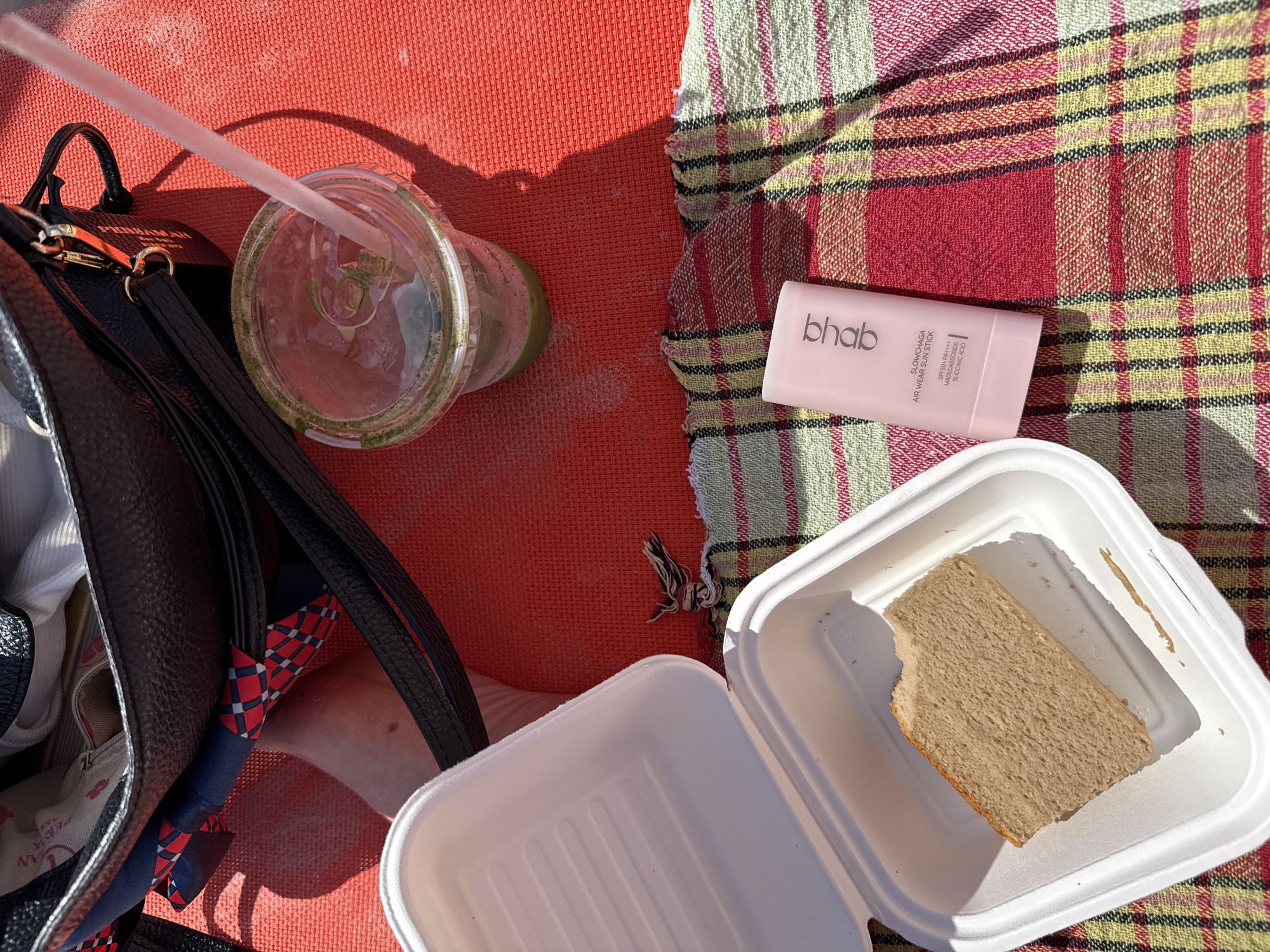 The image shows a picnic scene on a red textured mat, possibly in a park or beach setting in Tel Aviv, Israel, given the context of a sunny outdoor environment. A black leather handbag is placed on the mat, partially open, revealing some contents including a scarf with a red, blue, and white plaid pattern.
In front of the handbag, a clear plastic cup contains a green beverage, likely a smoothie or juice, with a light pink straw inserted. To the left of the cup are two white disposable food containers. The upper container is open and empty, while the lower container holds a single slice of light brown bread, with a small bite taken from it.
Below the food containers, a pink tube-shaped object, identified as "bhab SLOWCHAGA AIR WEAR SUN STICK SPF50+ PA++++ MADECASSOSIDE SUCCINIC ACID," lies on a checkered fabric cloth with red, green, yellow, and white stripes. The sunlight casts shadows on the scene, indicating it is daytime. The overall impression is one of a casual outdoor meal or snack.