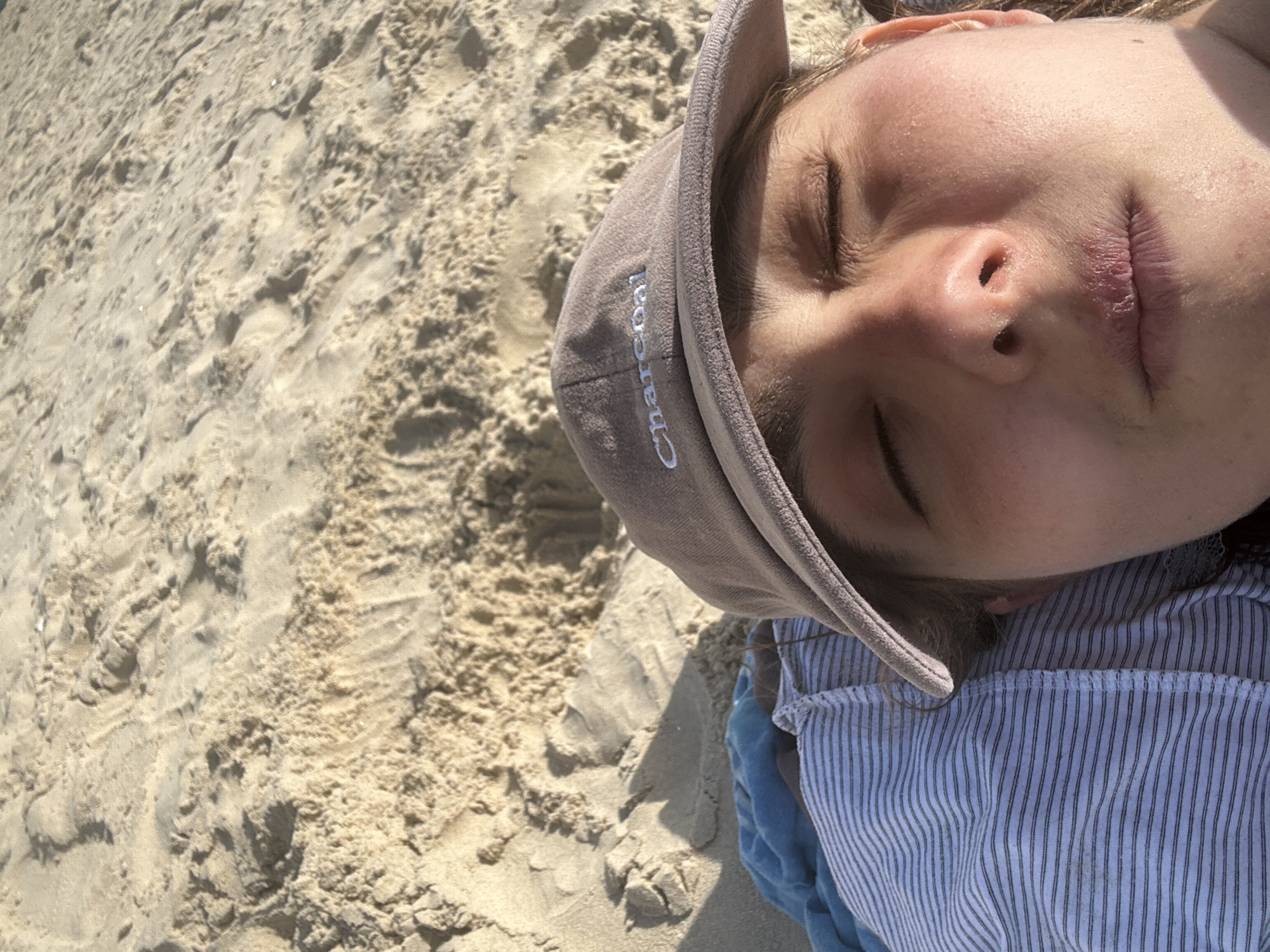 A child wearing a cap and striped shirt is lying on sandy terrain. The cap has the word "Charcoal" embroidered on its side. The child has their eyes closed. The sand has uneven texture, appearing to be wet in some areas. The location is presumed to be Tel-Aviv, Israel, based on the prompt. There are no discernible actions or interactions.