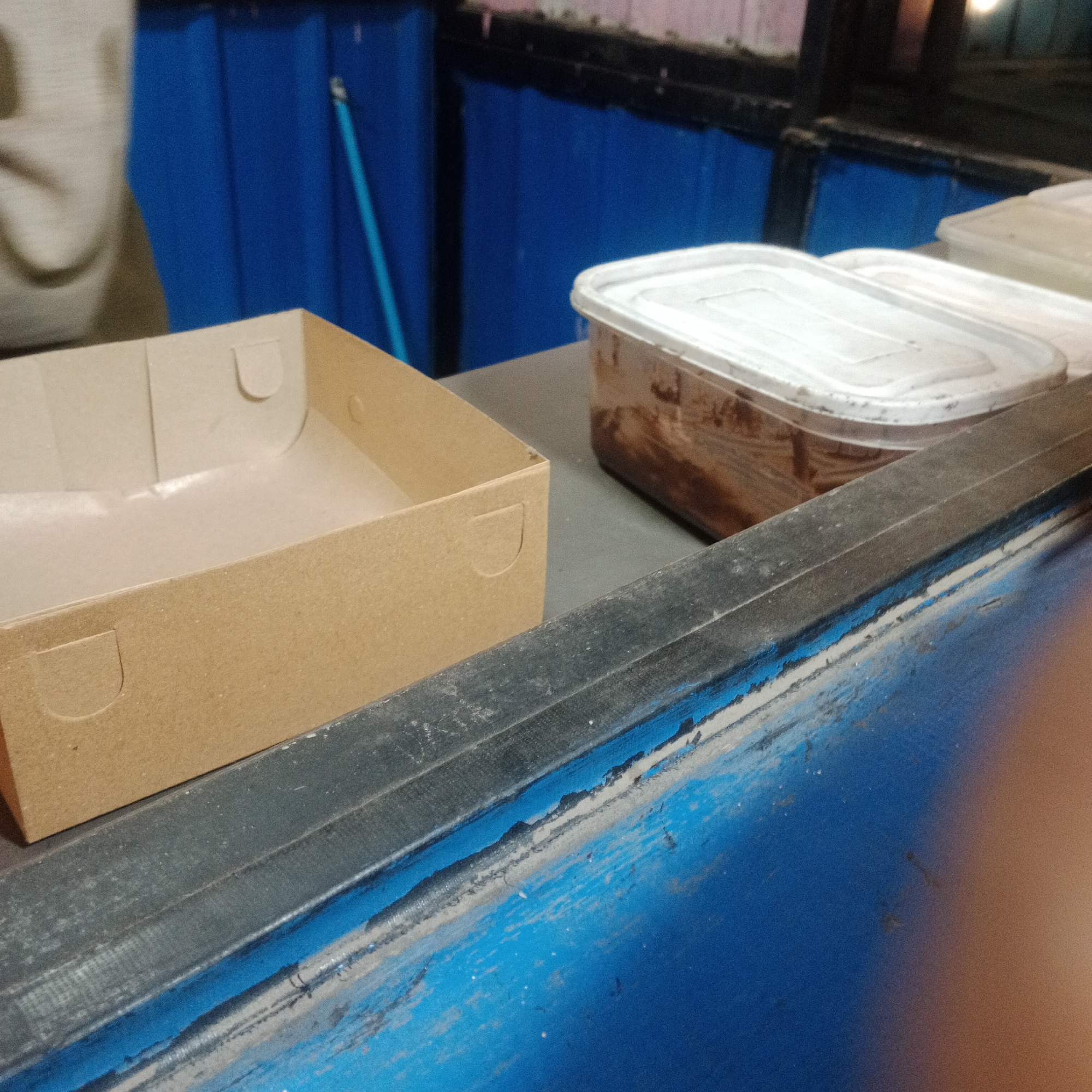 A brown cardboard box is in the foreground on the left, with a white plastic container of food on the right. A blue metal counter separates the foreground from the background, where a blue curtain is visible, and a person is partially visible on the far left. The scene appears to be in an outdoor food stall in Tegowanu Kulon, Indonesia, possibly during the day given the lighting, although the overall image is dark and grainy.