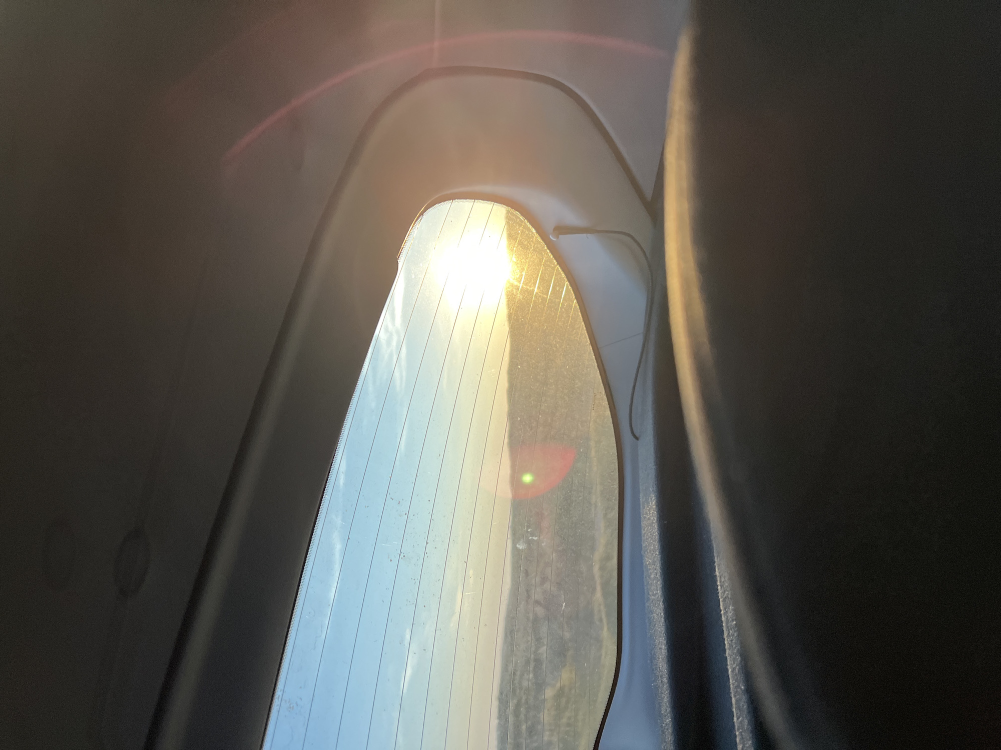 The image shows a view through the rear window of a vehicle, looking out at a rural landscape. The sun is setting, casting a warm glow and creating lens flare on the window. The landscape appears to be rolling hills with some vegetation. The interior of the vehicle, including a dark upholstered surface and the car's headliner, is visible in the foreground and upper part of the image. The image was taken in or near Carrickboyle, Ireland.