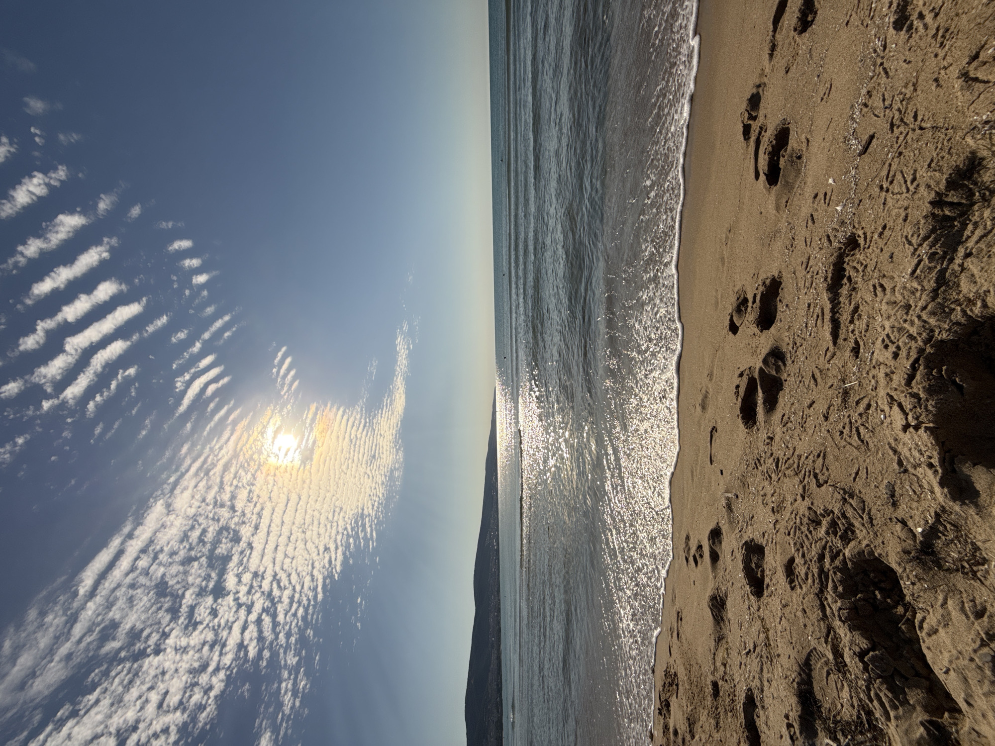 The image depicts a tranquil beach scene in Nesebar, Bulgaria. In the foreground, a sandy beach is covered with footprints and small debris. The gentle waves of the Black Sea are rolling onto the shore, creating a foamy white line. The sun is shining brightly, casting a shimmering reflection on the water. In the distance, a hilly coastline can be seen, with a clear blue sky overhead. The sky is partially covered by scattered clouds that form a distinctive pattern, with some appearing as parallel lines. The overall impression is one of a peaceful and serene day at the seaside.