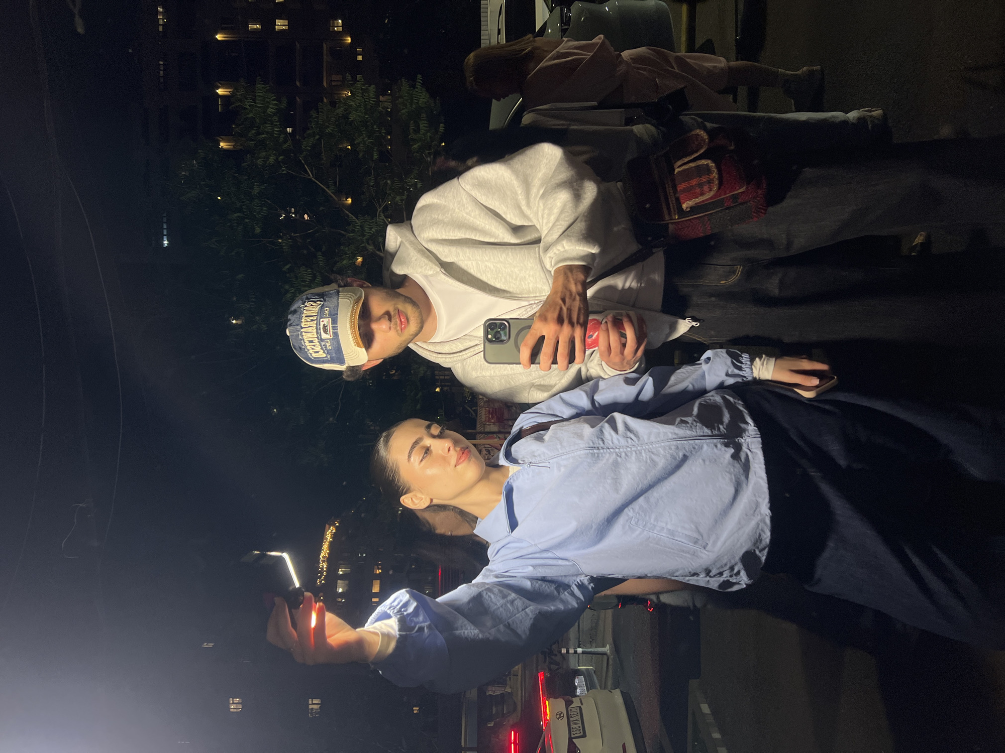 A man and woman stand in front of a parked white vehicle with red tail lights. The woman holds a phone, illuminating her face with the screen's light, and appears to be taking a selfie or a photo with the man. The man stands behind her, holding his own phone, possibly monitoring the photo. They are outdoors at night in an urban setting. Buildings are visible in the background. Trees are also visible. The license plate on the vehicle reads "859 MM 39". The precise location is not evident from the image, and therefore its location in the city of Yerevan, Armenia, cannot be confirmed.