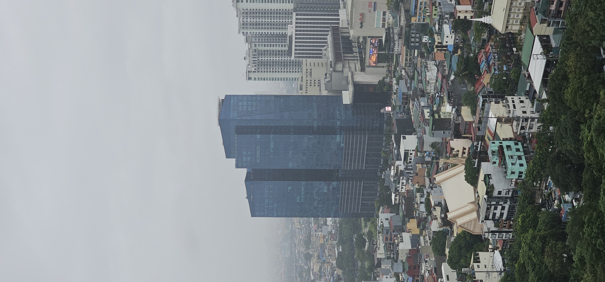 The image presents an aerial view of a dense urban landscape in Makati, Philippines, under an overcast sky. Dominating the scene are two large, modern skyscrapers with reflective blue glass facades, positioned centrally. To their right, a cluster of residential buildings with varying architectural styles and colors are visible, some featuring balconies and prominent air conditioning units.
Further into the foreground, a striking white church with a distinctive pointed roof and a spire stands out. Surrounding the church are lower-rise buildings, many with brightly colored roofs and weathered facades, indicative of a mixed residential and commercial area. Dense foliage, primarily trees, occupies the lower portion of the frame, suggesting a park or a residential area with abundant greenery. In the distance, a hazy cityscape stretches to the horizon, with numerous buildings of different heights and architectural designs, characteristic of a sprawling metropolis. The overall impression is one of urban density and a blend of modern and older architectural styles.