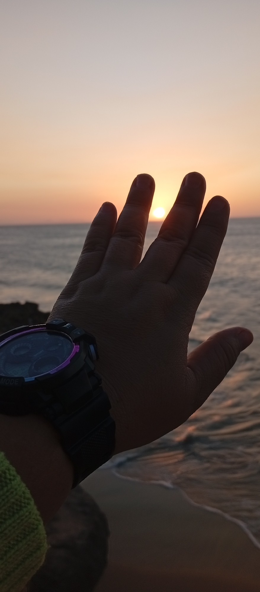 A person wearing a digital watch on their left wrist holds their hand up towards the setting sun over the ocean. The hand partially obscures the sun, which is visible between the fingers. The location appears to be near a beach, possibly in or near Juan Griego, Venezuela, due to the coastal setting and sunset. Waves can be seen on the beach. The lighting is soft due to the sunset.