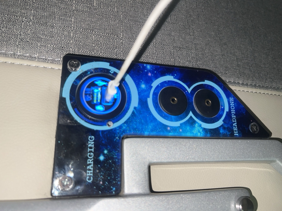 The image displays an integrated multi-function panel within an interior environment, located in Aê Thi, Vietnam. The panel features a dark blue surface with a starfield pattern, secured by four visible screws.
The upper portion of the panel is labeled "CHARGING" in white text. Within this section, a circular module emits a bright blue light, indicating active function. This module contains two stacked USB-A ports, one of which has a white USB-A cable inserted, glowing with blue light at the connection point. The visible label "QC 3.0" indicates Quick Charge 3.0 capability for the USB ports.
The lower portion of the panel is labeled "HEADPHONE" in white text and contains two vertically arranged 3.5mm audio jacks. These jacks are black with visible gold-colored internal contacts.
The panel is flanked by structural and upholstered elements. To its left, a light grey, textured frame or support structure is visible, behind which a segment of cream-colored upholstery is present. To its right, a grey, tightly woven fabric, consistent with seat upholstery, is visible. No individuals are depicted in the scene.