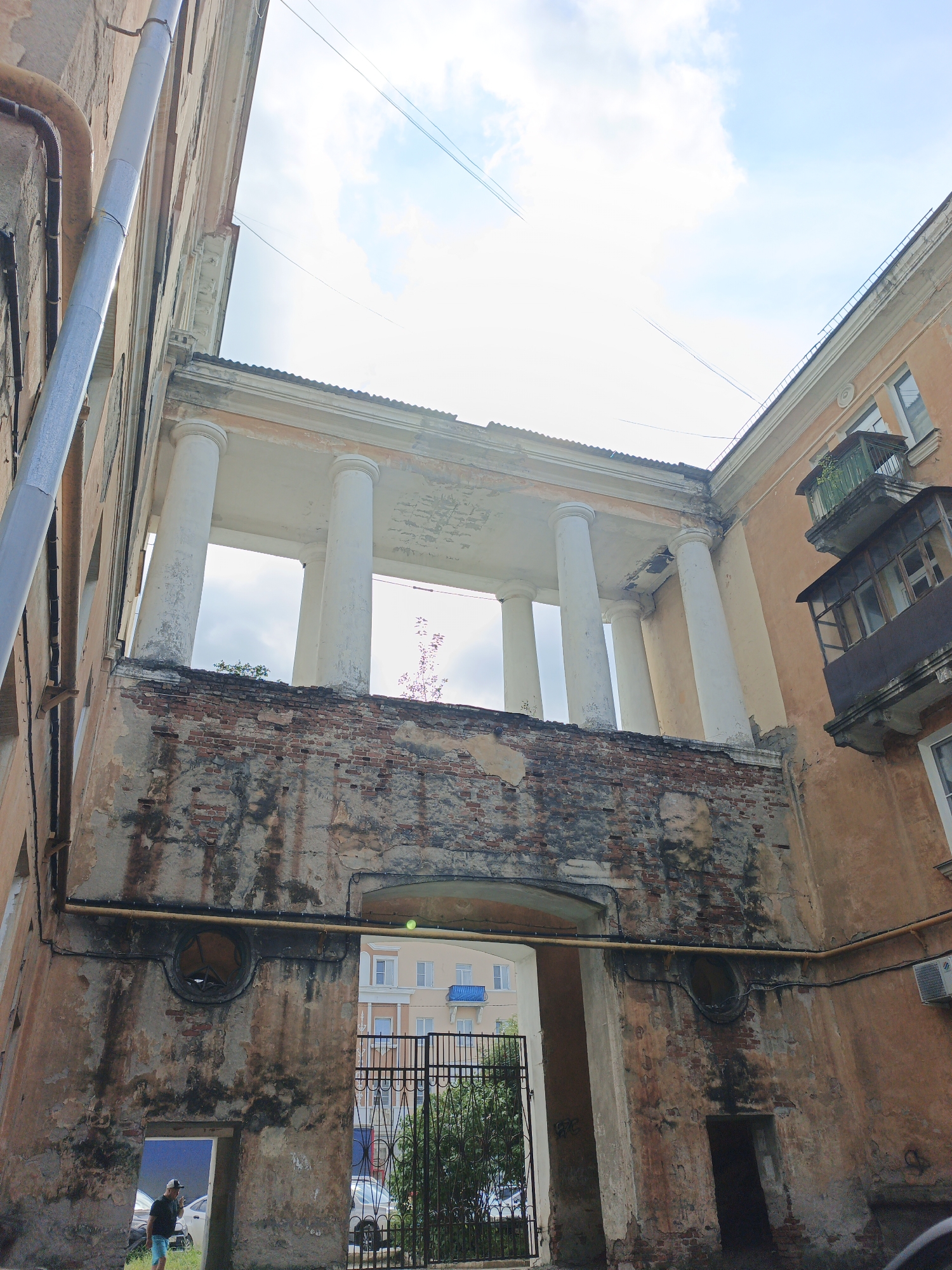 An exterior low-angle shot captures a dilapidated, orange-hued building with visible signs of decay. A prominent feature is a colonnade with at least four large, white, fluted columns supporting a partial roof structure. Below the colonnade, a section of brick wall has crumbled, exposing the underlying red bricks and mortar. Archways are visible, one of which leads to a courtyard with a black metal gate and greenery. Another archway appears to be an entrance to a darkened interior space. A metal pipe runs horizontally across the facade. To the right, balconies with rusty railings and glass panes extend from the building. In the foreground, a person wearing a dark t-shirt, blue shorts, and a baseball cap stands near a doorway, looking to the left. Cars are visible in the background. The sky above is bright blue with scattered white clouds and thin contrails. The building appears to be located in Zlatoust, Russia.