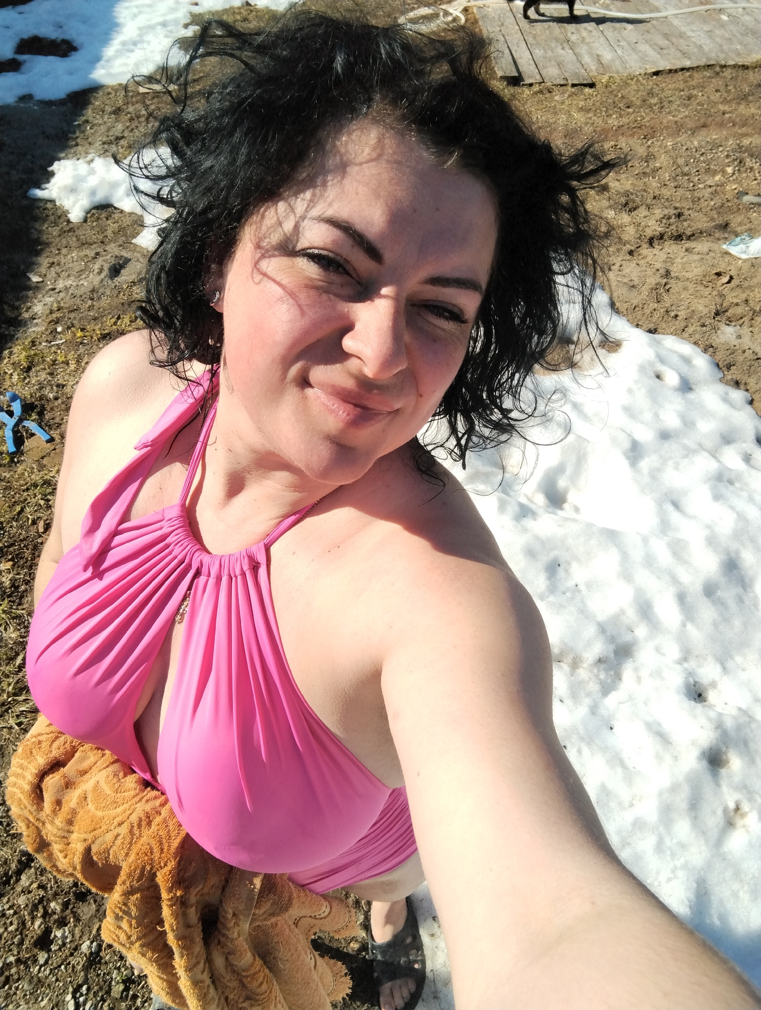 A woman with dark, wet curly hair poses in a pink swimsuit against a backdrop of melting snow and mud. She is holding the camera at arm's length, taking a selfie. A brown towel is draped over her. Patches of snow are visible on the ground. In the background, there is a wooden structure with a dark object that could be an animal, possibly a cat. Blue pliers are on the ground. The exact location in the city of Grishanki, Russia cannot be determined from the image alone.