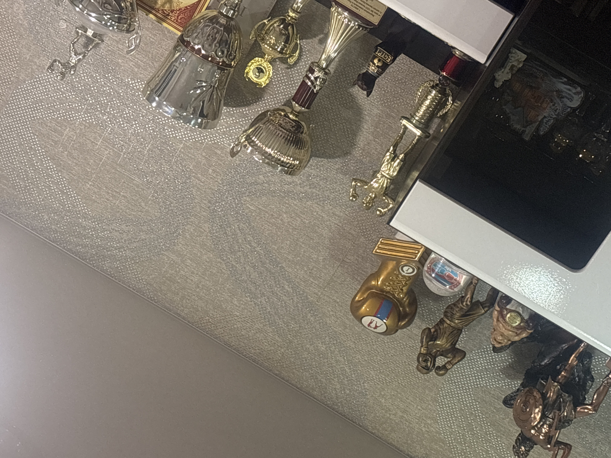 The image depicts a collection of trophies and figurines displayed on a shelf against a textured, light-colored wall. On the left side of the shelf, a large bronze figurine of a warrior with a shield and spear is visible. Next to it, a smaller, portly figure wearing a medal is placed. Further to the right, a bronze figurine of a boxer in mid-action stands. Beside the boxer figurine is a golden trophy shaped like a boxing glove with blue and red stripes on it.
The middle section of the shelf features a golden trophy with a detailed cup and a dark red stem. To its right, a small black figurine, possibly a cat or an ankh symbol, is adorned with a golden collar that reads "EGYPT". Adjacent to this is a smaller golden trophy with a circular emblem. To the far right, two silver trophies are displayed, one larger with ornate detailing and a red band, and another slightly smaller.
The shelf itself appears to be white and white-bordered. Reflections in a dark surface on the left of the image suggest the presence of a cabinet or display case. The overall scene suggests a home or personal space in Novinki, Russia, where these awards and collectibles are showcased.