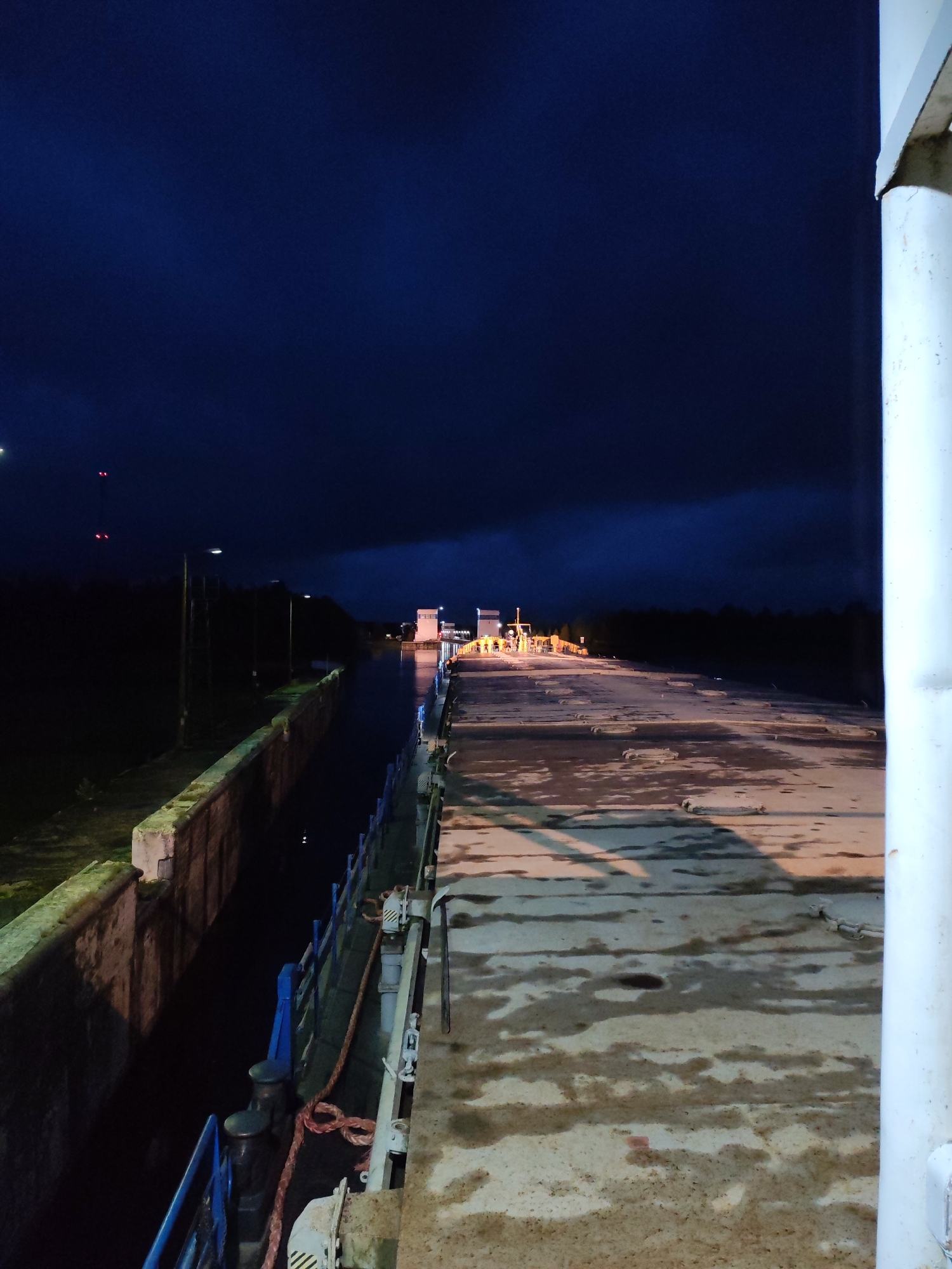 A nighttime shot depicts the exterior of a lock, likely part of a canal system, in the village of Devyatiny, Russia. The scene is illuminated by artificial lights, casting shadows and highlighting the wet, industrial surface of what appears to be a barge or lock gate. The water within the lock is dark and still. In the distance, illuminated structures suggest operational equipment, possibly part of the lock's machinery or a nearby building. The sky is a deep, dark blue, indicating night. The environment is characterized by concrete structures, metal railings, and bollards, consistent with a functional waterway. No individuals are clearly visible in the frame.