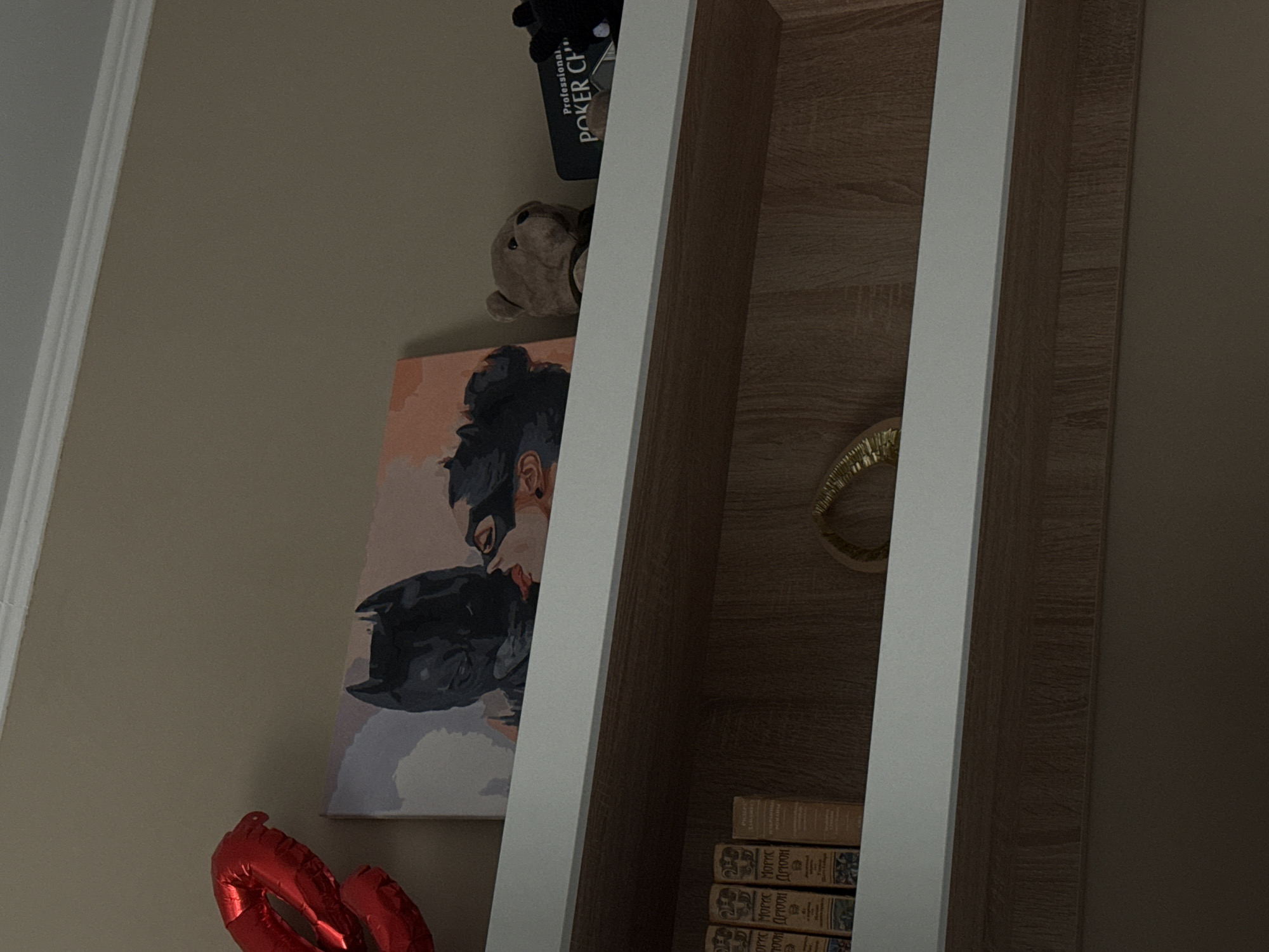 The image shows a section of a shelving unit containing various objects. A painting depicting a woman with dark hair and a mask is visible, along with a teddy bear, a "Professional Poker Chip" box, a gold bracelet, and stacked books. A red balloon is positioned below the painting. The shelves have a light wood-grain finish with white framing. The location and city are not identifiable from the image.