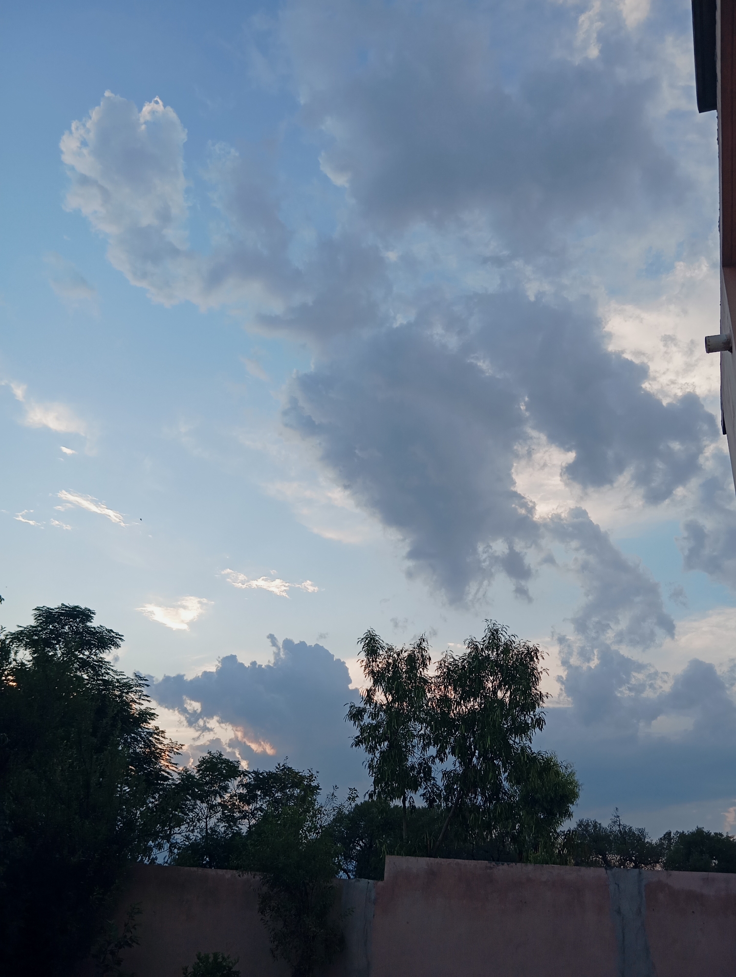 An outdoor, low-angle, full shot captures a wall and trees with a cloudy sky above. The wall, constructed from concrete blocks and possibly stucco, spans the foreground horizontally. Behind the wall, dense trees with dark green foliage are silhouetted against a sky filled with varying shades of blue and grey clouds. Some clouds are light and wispy, while others are dark and heavy, suggesting impending weather changes. A sliver of bright light is visible between some of the clouds in the middle ground, hinting at the setting sun. A small, dark object, possibly a bird, is visible against the sky in the upper left quadrant. On the far right, the edge of a building with a dark roofline and a white pipe extending from it is visible. The scene is devoid of people or any discernable activity.