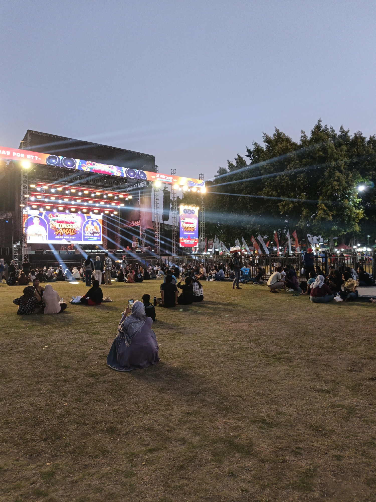 This image depicts an outdoor concert scene at night in Klaten, Indonesia. A large stage is set up with bright lights and screens displaying event information, including text that appears to be in Indonesian. A crowd of people are seated on a grassy field in front of the stage, facing the performance. Some individuals are taking pictures with their phones, while others are socializing. The overall atmosphere suggests a community event. The presence of Indonesian text on the stage screens indicates the specific location within Indonesia.