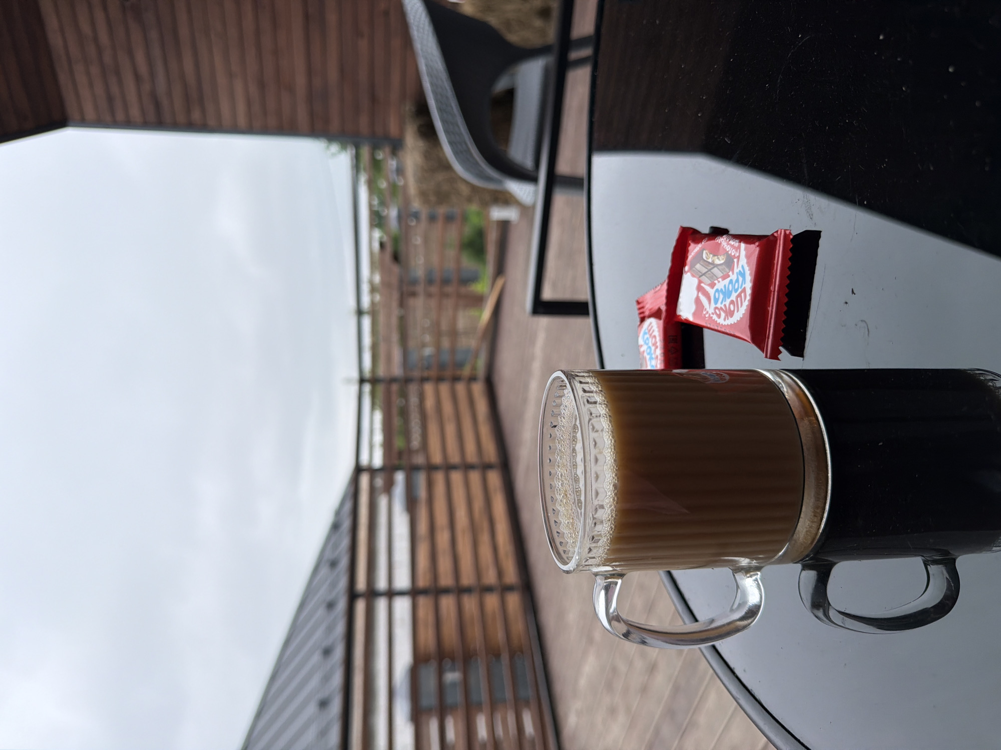 A glass mug filled with coffee sits on a black reflective table. Two red candy wrappers are next to the mug. The table is on a wooden deck, which appears to be part of a porch or patio. In the background, a wooden railing separates the deck from a sloped roof structure. Beyond the railing, there's a cloudy sky and some greenery. The scene is likely outdoors, on the porch of a house or cottage, possibly in the city of Semiluki, Russia, given the architectural style and natural surroundings.