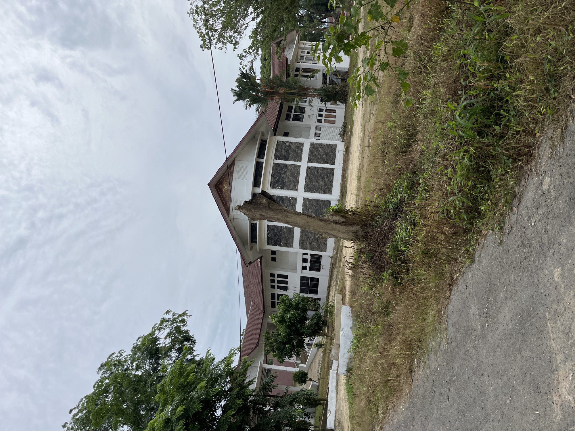 The image shows a large, multi-story building with a white facade and a dark red tiled roof. The building has several windows and appears to be constructed with stone accents. In the foreground, there is a dirt road and overgrown vegetation. A dead tree trunk stands prominently in the middle ground. The sky is overcast. The building is likely located in a suburban or rural area of Bundar, Indonesia, given the surrounding vegetation and unpaved road.