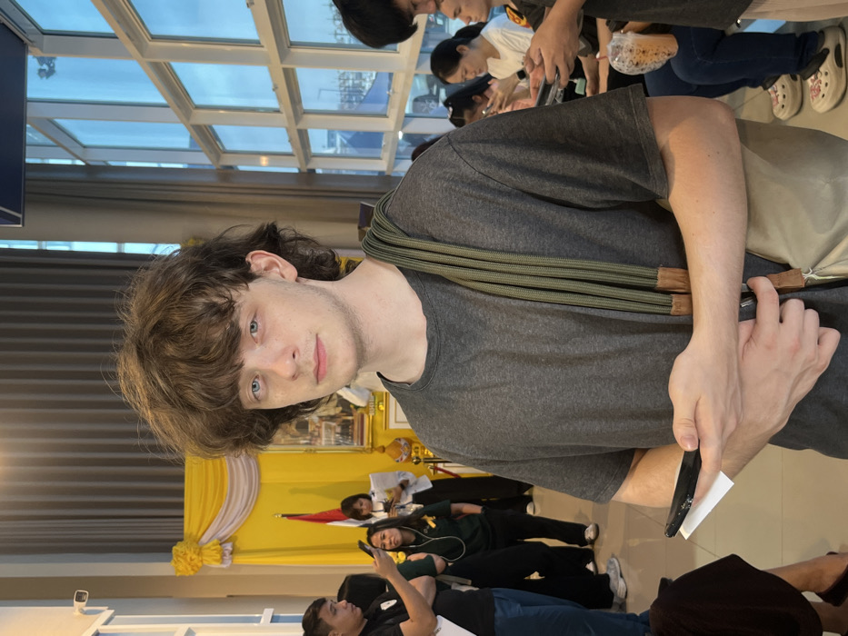 The image depicts an indoor scene within a public building in Samut Prakan, Thailand. A young Caucasian male with medium-length brown hair and blue eyes stands prominently in the foreground, wearing a dark grey t-shirt. An olive green multi-strand cord is draped over his left shoulder, connected to a beige bag. He holds a black smartphone horizontally in his left hand, with a white paper slip tucked beneath it, while his right hand clasps his left forearm. He gazes directly forward with a neutral expression.
To the subject's left, a formal display features yellow and white fabric draping, a red and white striped flag, and a framed portrait, possibly of a revered figure, consistent with cultural decor in Thailand. Several individuals are visible in this background area; one in a black polo shirt operates a smartphone, and another in a white shirt holds papers. A white security camera is mounted high on the wall.
To the subject's right, large windows reveal an exterior view of a body of water with numerous boats moored, suggesting a harbor or riverfront setting, typical for coastal Samut Prakan. The sky is clear blue. Several individuals are seated along a wall or bench on the right, predominantly engaged with their mobile phones. One individual has a plastic cup containing a brown beverage, and is wearing white decorated clogs. The interior flooring consists of light-colored tiles.