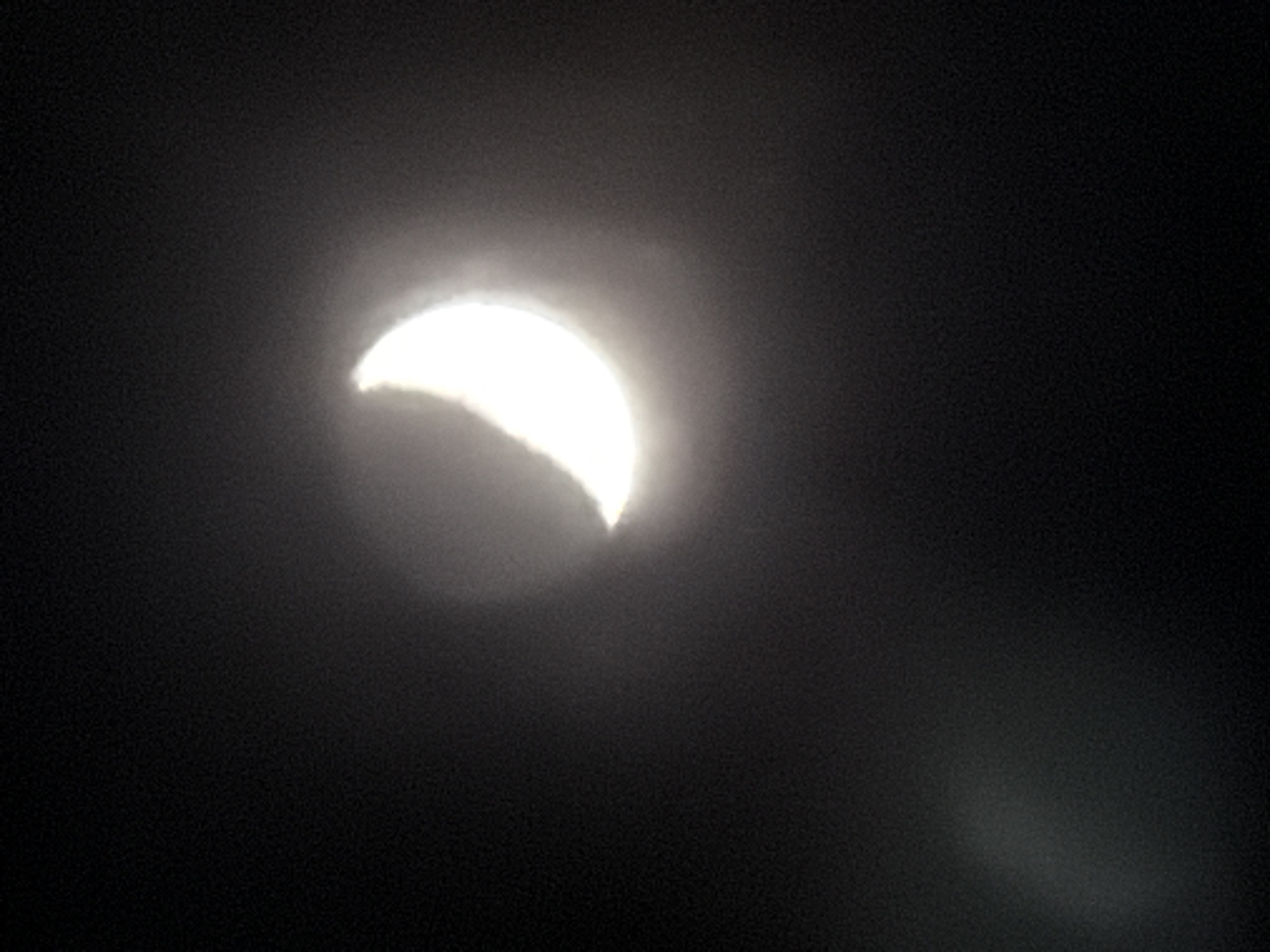 The image captures a celestial event featuring a brilliant, crescent-shaped light source against a dark background, strongly suggesting a partial solar eclipse. The bright crescent, intensely white and glowing, is positioned in the upper-left portion of the frame, emitting a soft, diffused halo of light that blurs its edges. This diffusion indicates the presence of atmospheric conditions such as haze, mist, or thin clouds scattering the light. The surrounding environment is uniformly dark, creating a stark contrast with the luminous crescent. The clean, curved line delineating the 'missing' portion of the celestial body is characteristic of an eclipse. No surface details are discernible on the bright crescent due to its overwhelming luminosity and the atmospheric effects. There are no visible people, other objects, or text. The scene appears to be set during daytime, with the dark background likely a result of exposure settings focused on the extreme brightness of the sun, or specific viewing conditions. The image conveys a powerful and ethereal natural phenomenon. Given the provided context, this event was observed in Markovo, Russia.