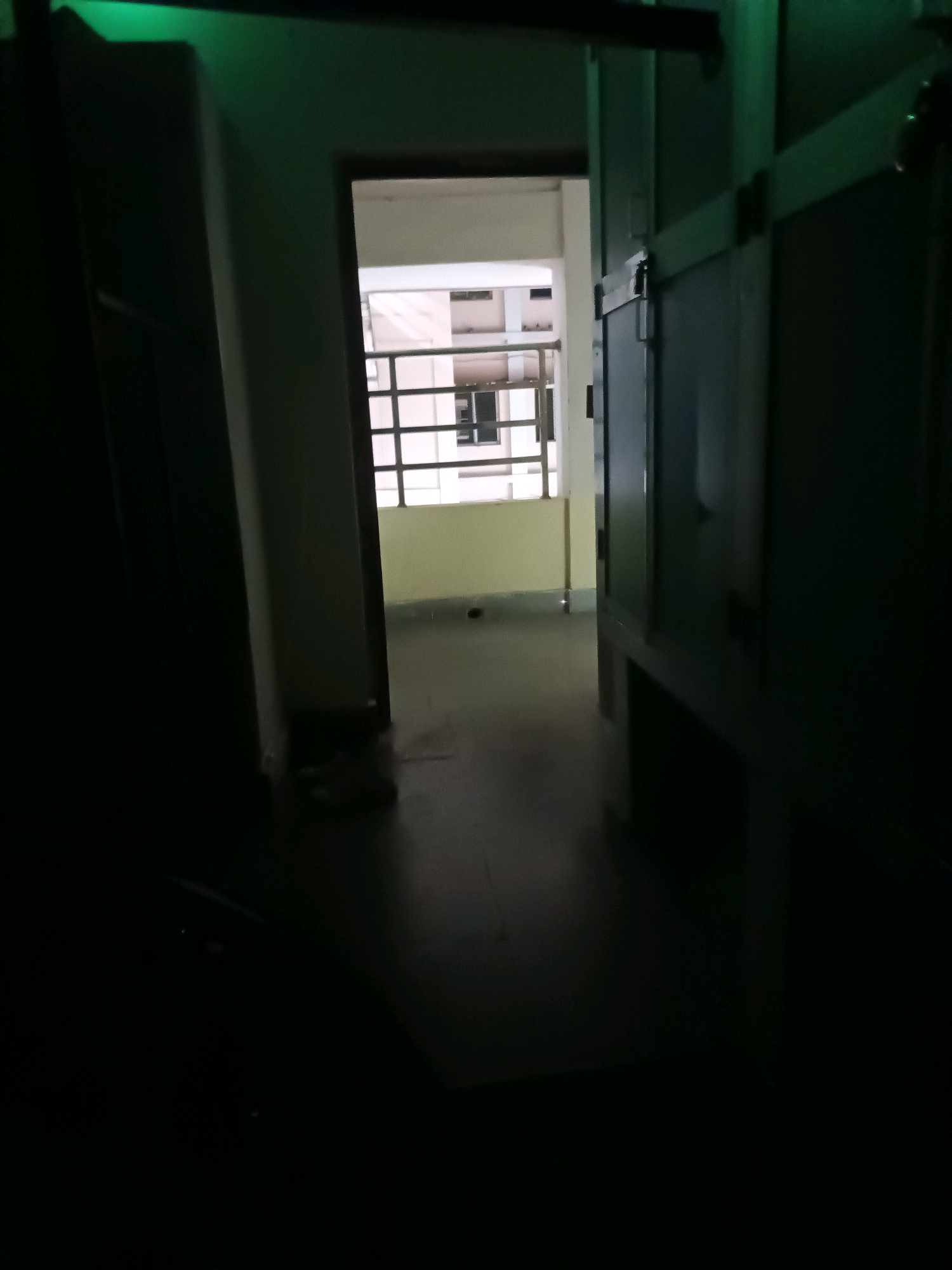 The image captures a dimly lit interior hallway. On the right side of the frame, a dark green door or cabinet with visible horizontal paneling and a metallic latch is present. To the left, a lighter colored wall extends towards the background. In the center, a doorway opens into another area which appears to be a room or corridor with a window. The window is divided into multiple panes by a grid-like structure and is partially illuminated. The floor in the hallway is a dark, matte surface. A green light is cast from above, illuminating the top portions of the walls and cabinets. The scene is devoid of any visible people or immediate activity, presenting a static, indoor environment. The location is specified as Sitalapalli, India.
