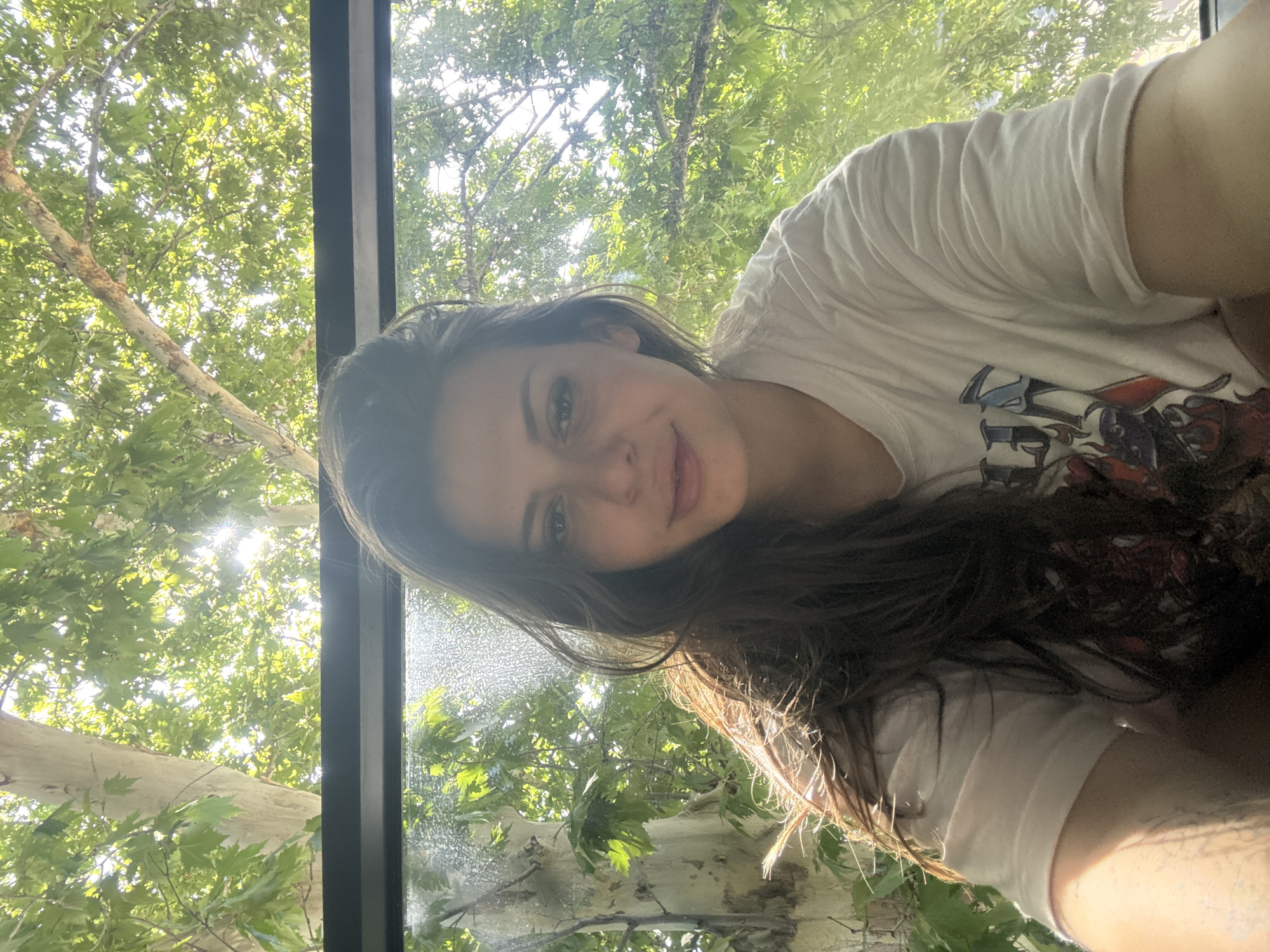 A young woman with long, dark hair is captured in a close-up selfie. She is wearing a white t-shirt with a graphic design. The background is dominated by lush green foliage and tree trunks, suggesting an outdoor setting with abundant natural light filtering through the leaves. The image appears to be taken during the daytime. The specific location in Plovdiv, Bulgaria cannot be determined from the visual information provided.